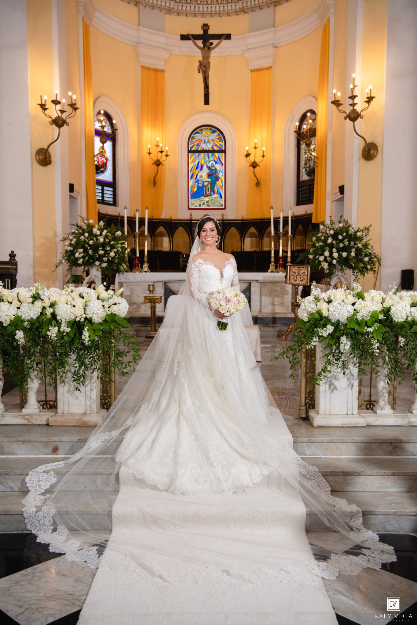 Bride in elegant white ball gown with long veil stands at church altar adorned with white floral arrangements.