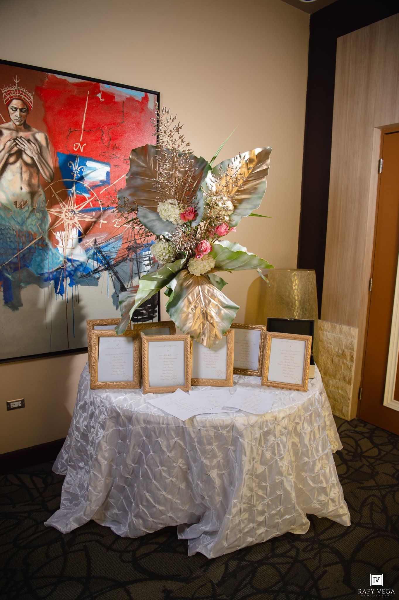 Elegant wedding seating chart table with gold frames, floral arrangement, and white linen before colorful artwork.