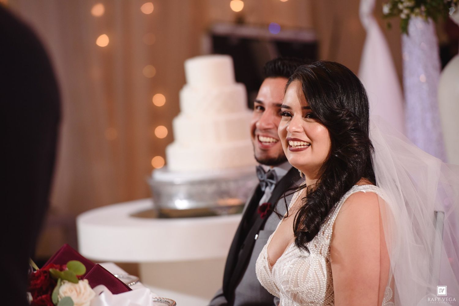 Smiling bride and groom at wedding reception with white tiered cake and warm fairy lights in background.