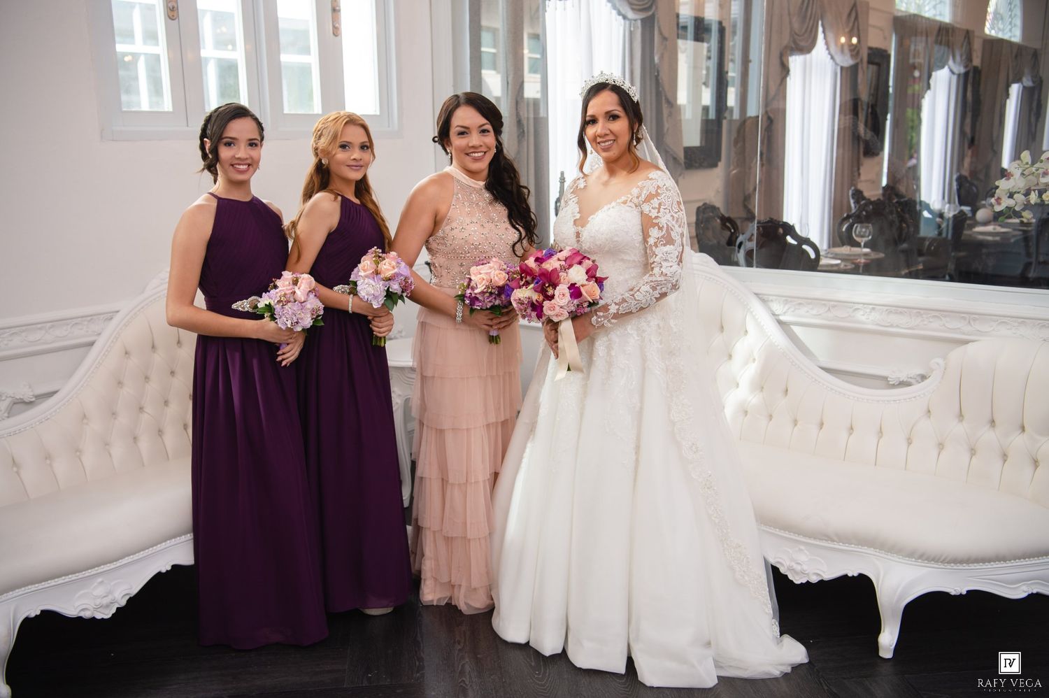 Bride in white gown poses with three bridesmaids in purple and blush dresses holding bouquets in elegant venue.