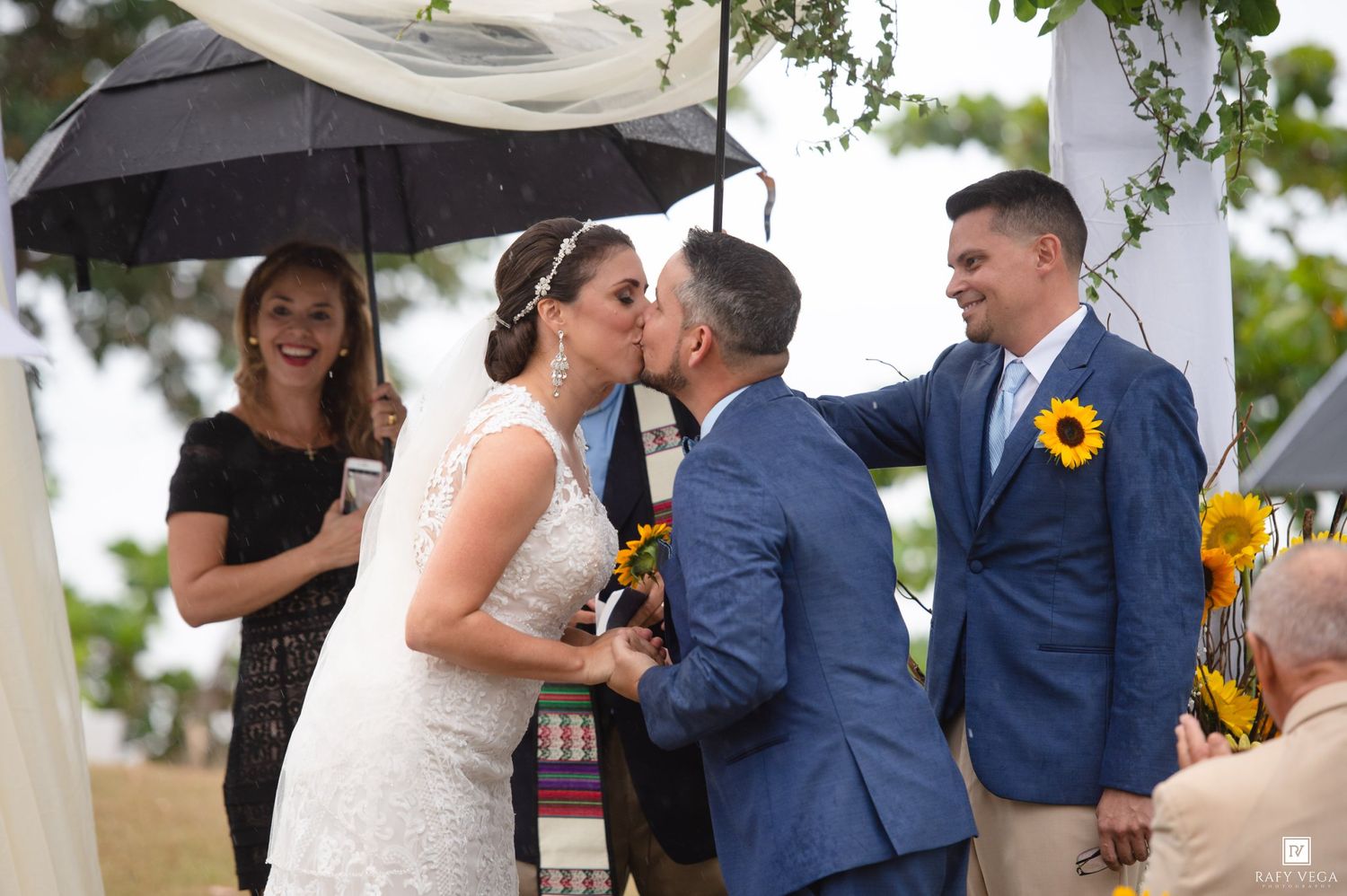 Bride and groom share a kiss at outdoor wedding ceremony with sunflowers, umbrella, and guests nearby.