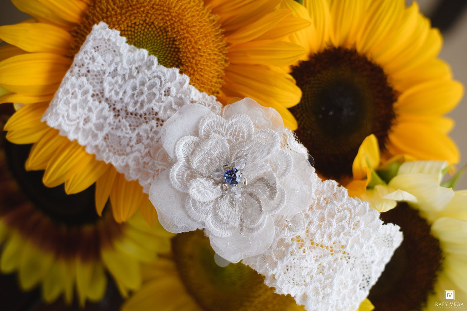 White lace bridal garter with floral detail and blue gem accent, displayed against vibrant yellow sunflowers.