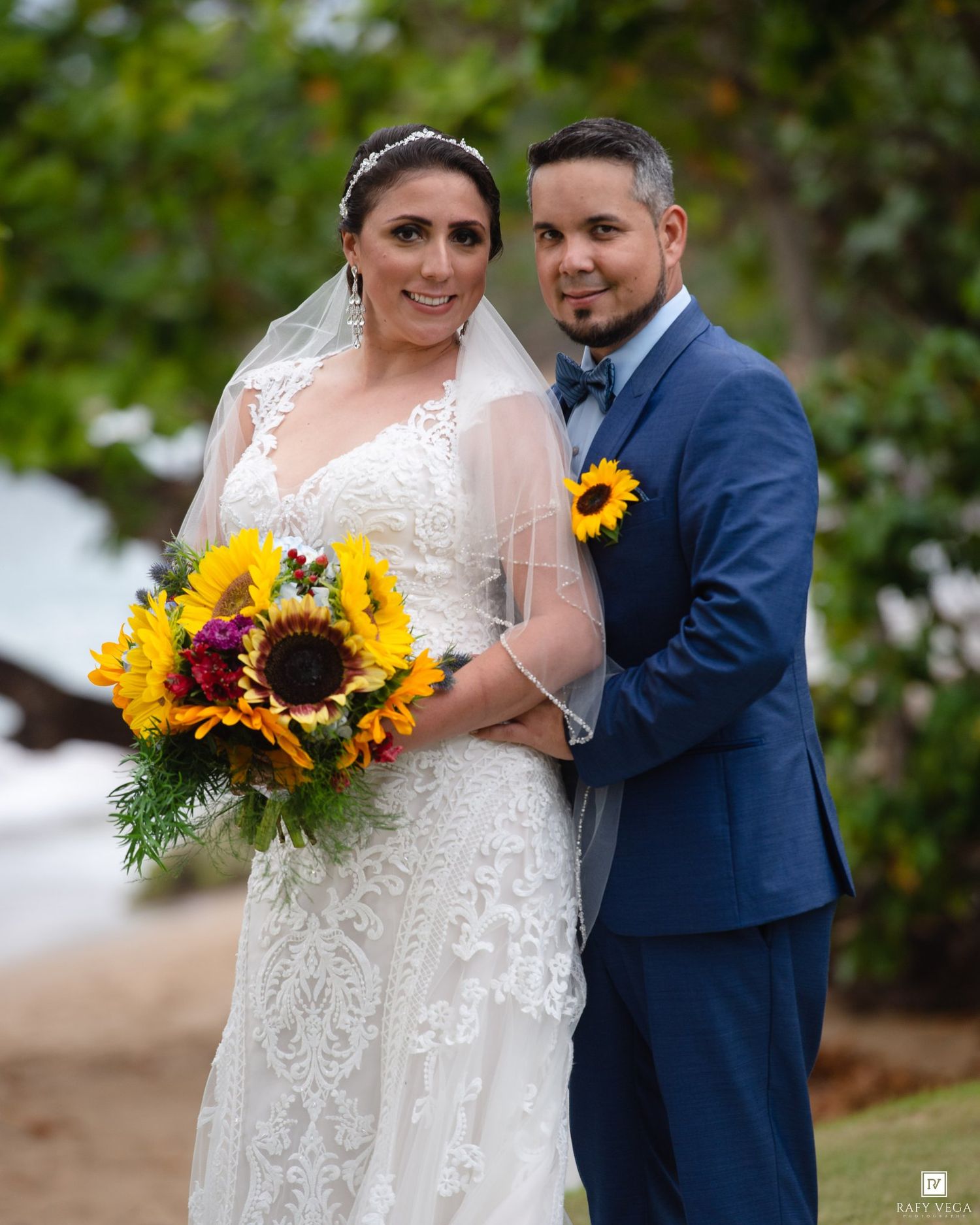 Bride in lace gown with sunflower bouquet and groom in blue suit pose for wedding portrait outdoors.