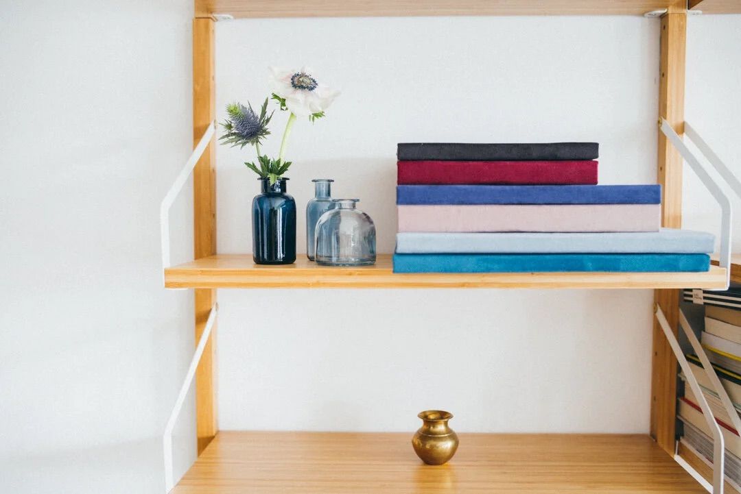 Wooden shelf with stacked colorful books, blue glass vases with flowers, and a small gold vase on white wall.