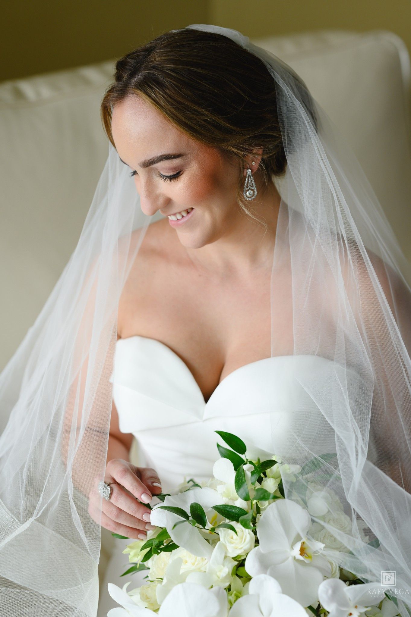 Smiling bride in strapless white gown and veil holds white orchid bouquet, wearing elegant drop earrings and diamond ring.