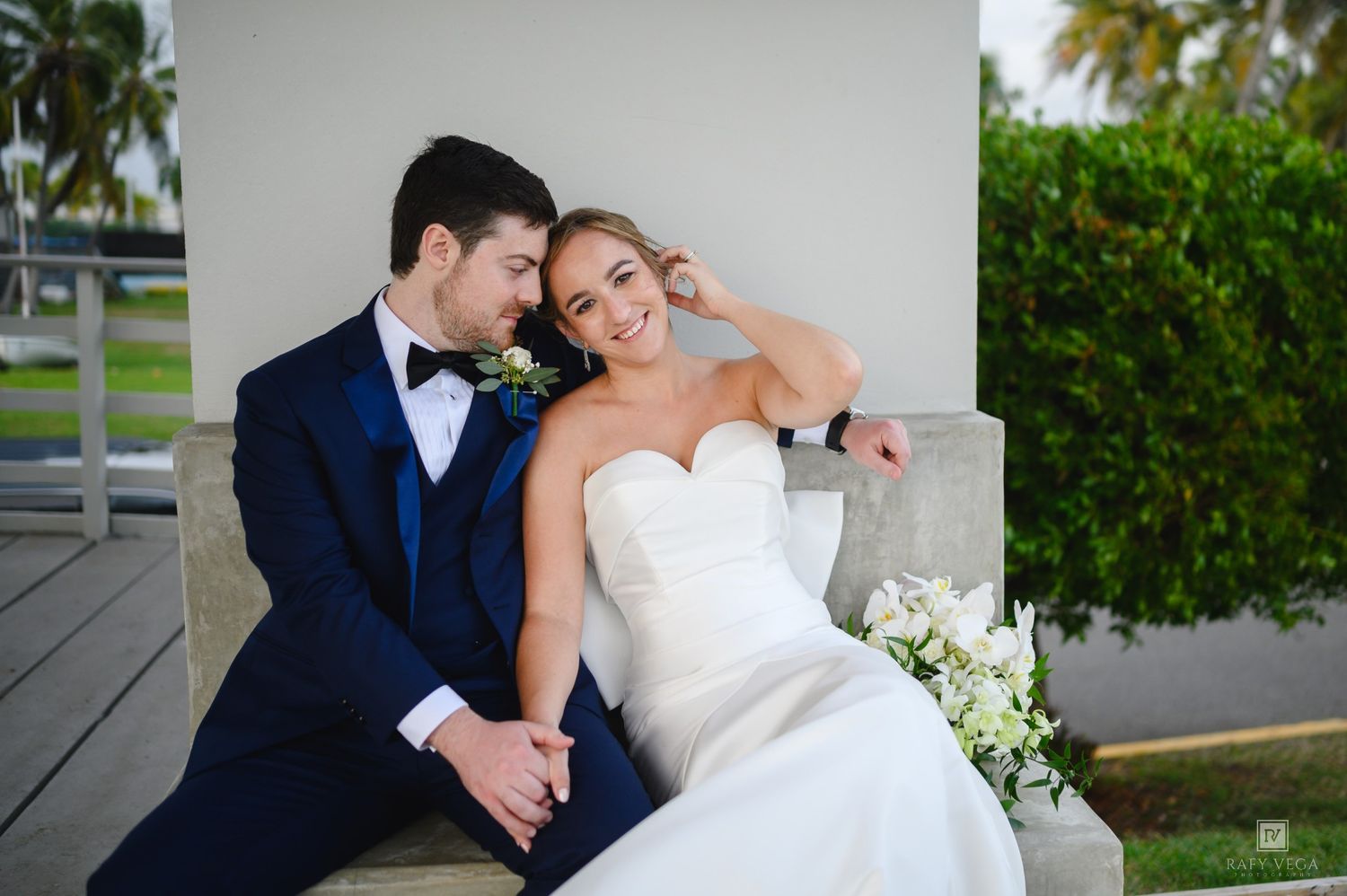 A bride and groom share a romantic moment seated outdoors, she in a strapless white gown, he in a navy blue tuxedo.