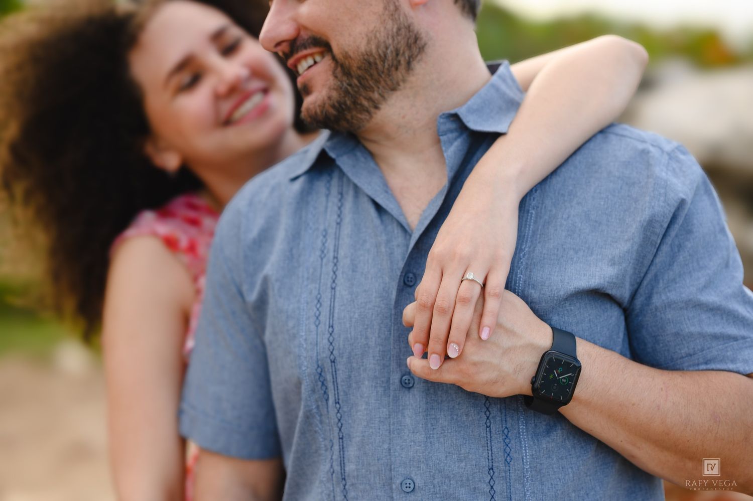 Couple embracing outdoors, woman showing engagement ring while smiling, man in blue shirt wearing a smartwatch.