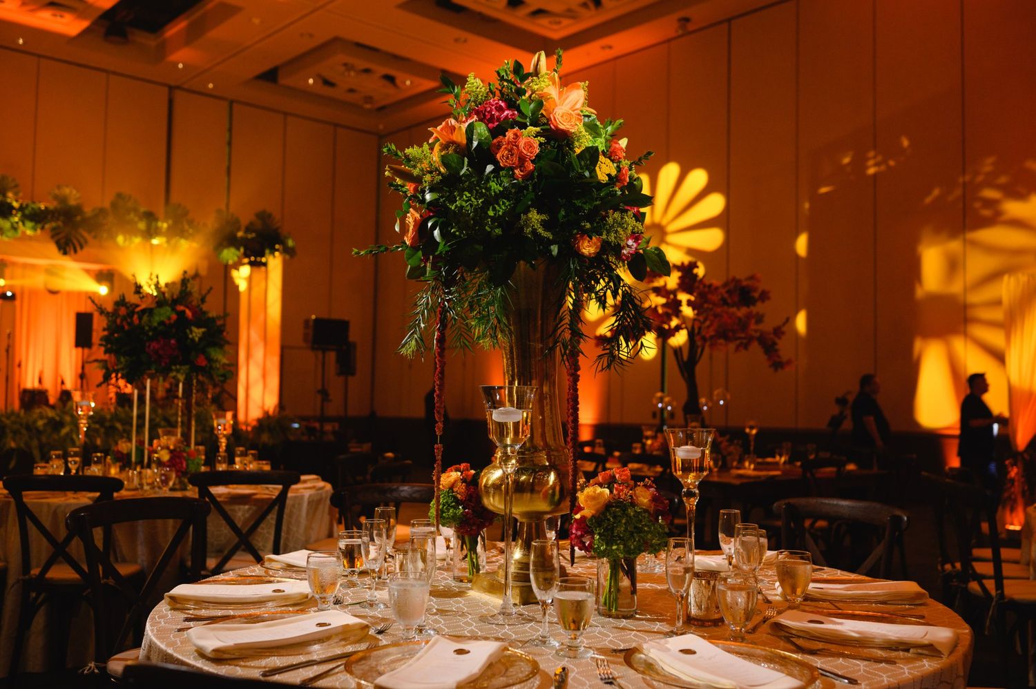 Elegant wedding reception with tall floral centerpieces, warm amber uplighting, and round tables set with glassware in a ballroom.