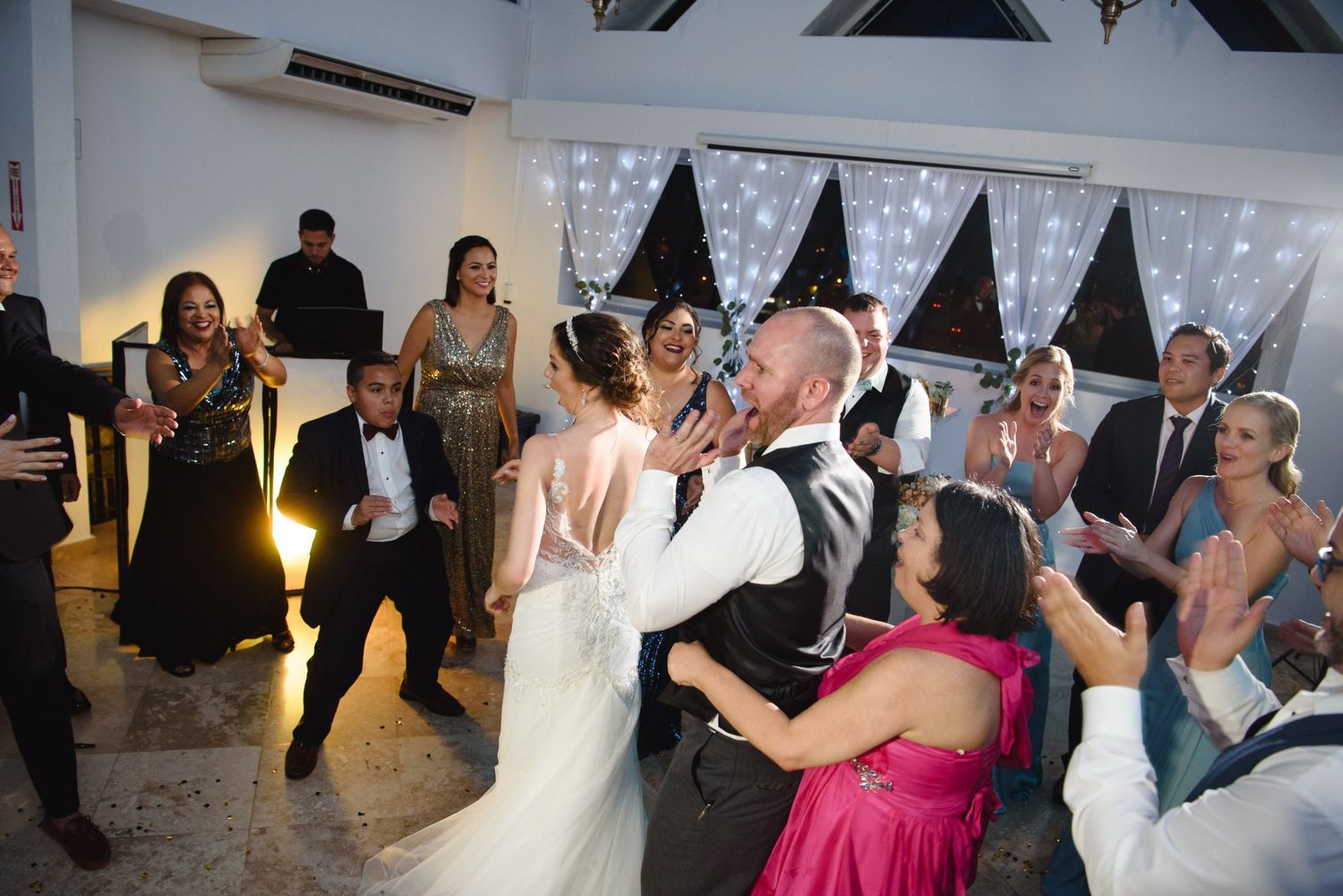 Bride and groom share a kiss on the dance floor surrounded by celebrating wedding guests in elegant attire.