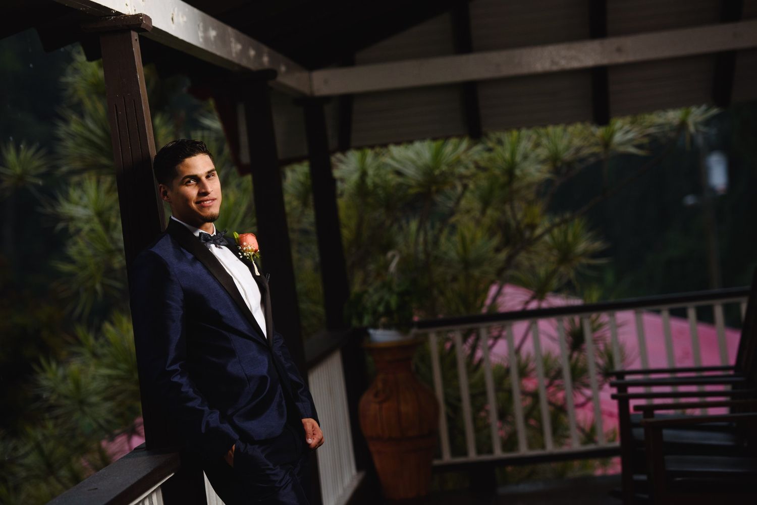 Groom in navy tuxedo with bow tie and boutonniere poses on a decorated porch at night.