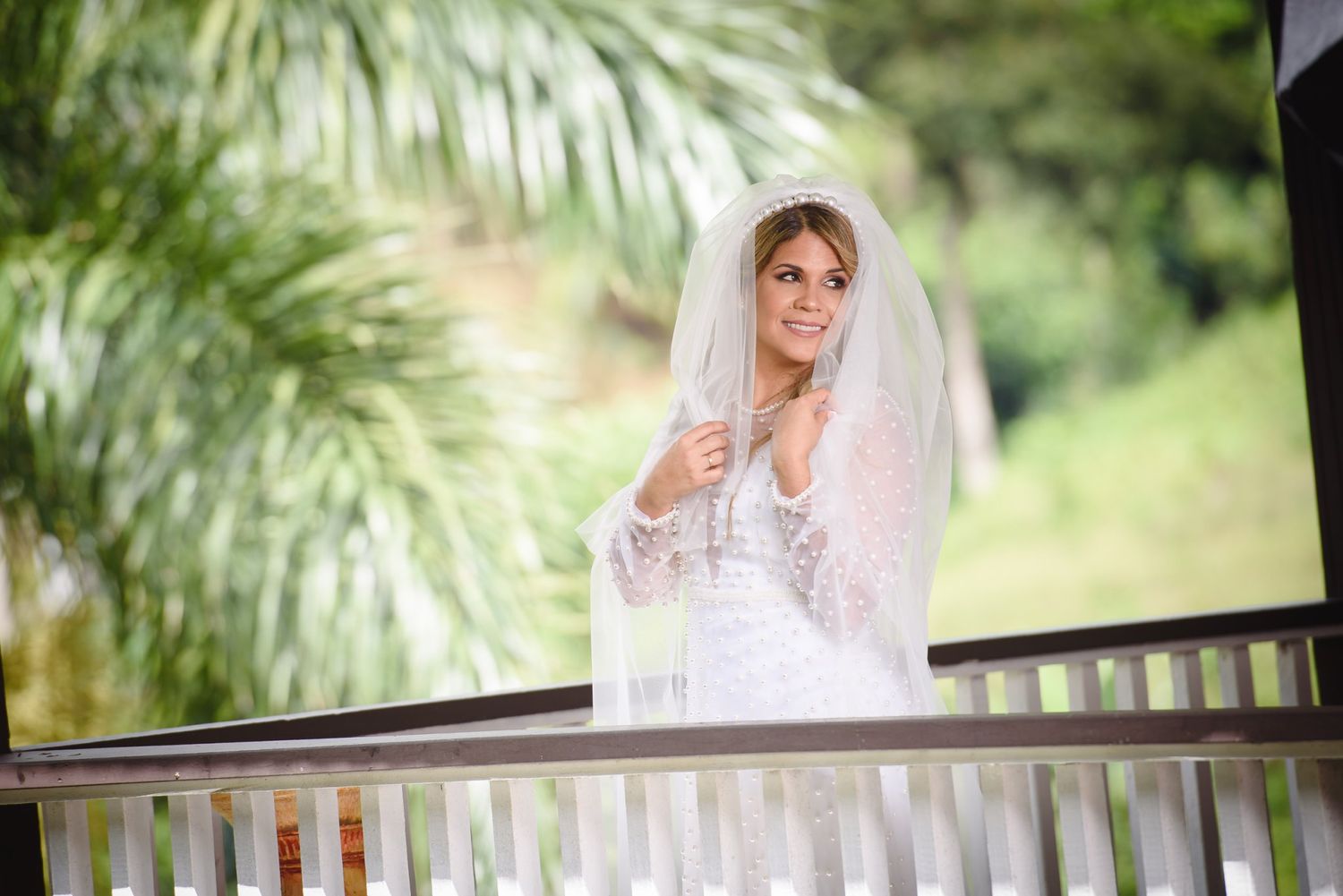 Bride in white lace wedding dress and veil standing on a balcony surrounded by lush green tropical foliage.