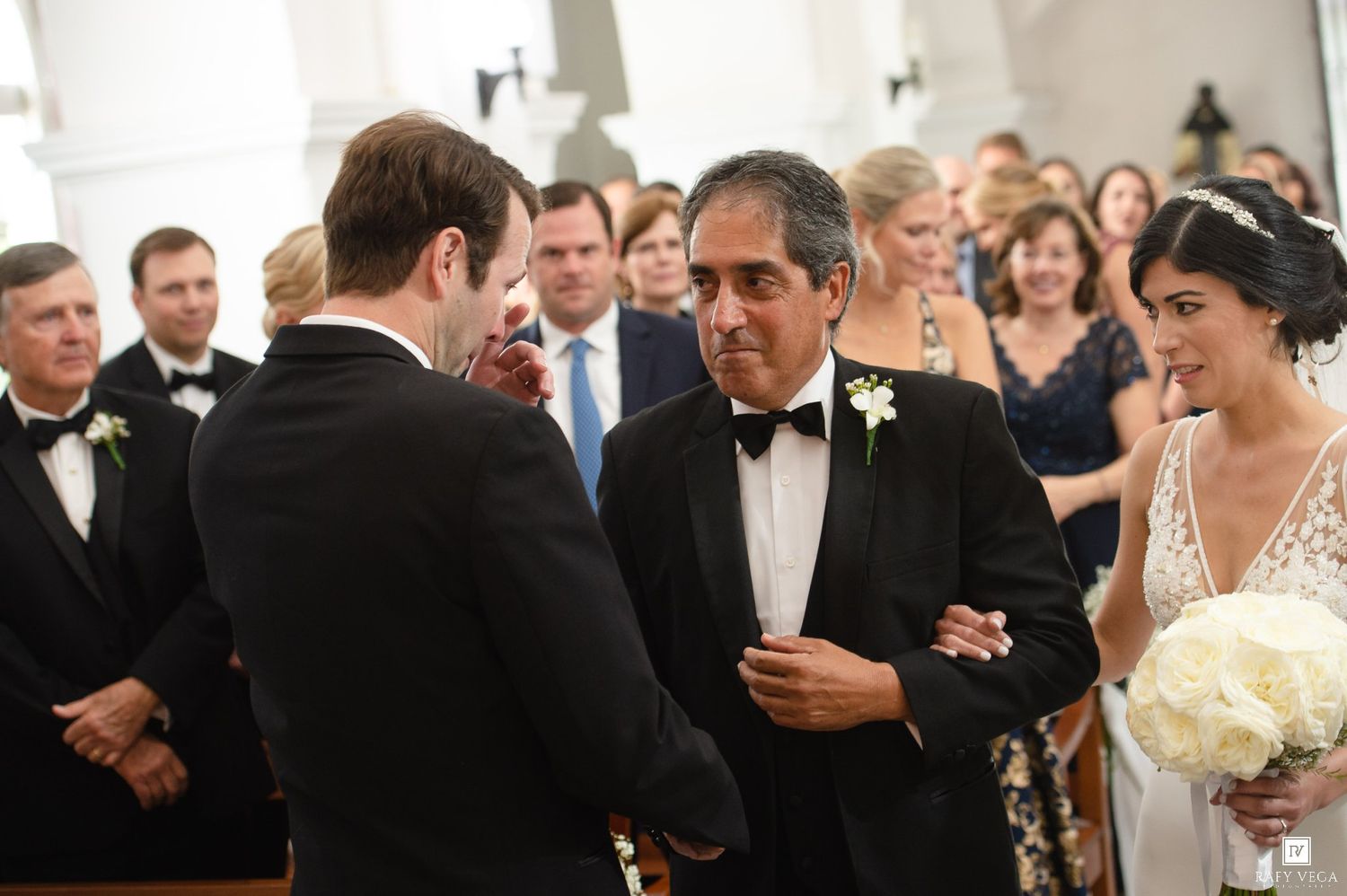 Father of the bride shaking hands with groom at church wedding ceremony, bride holding white bouquet.