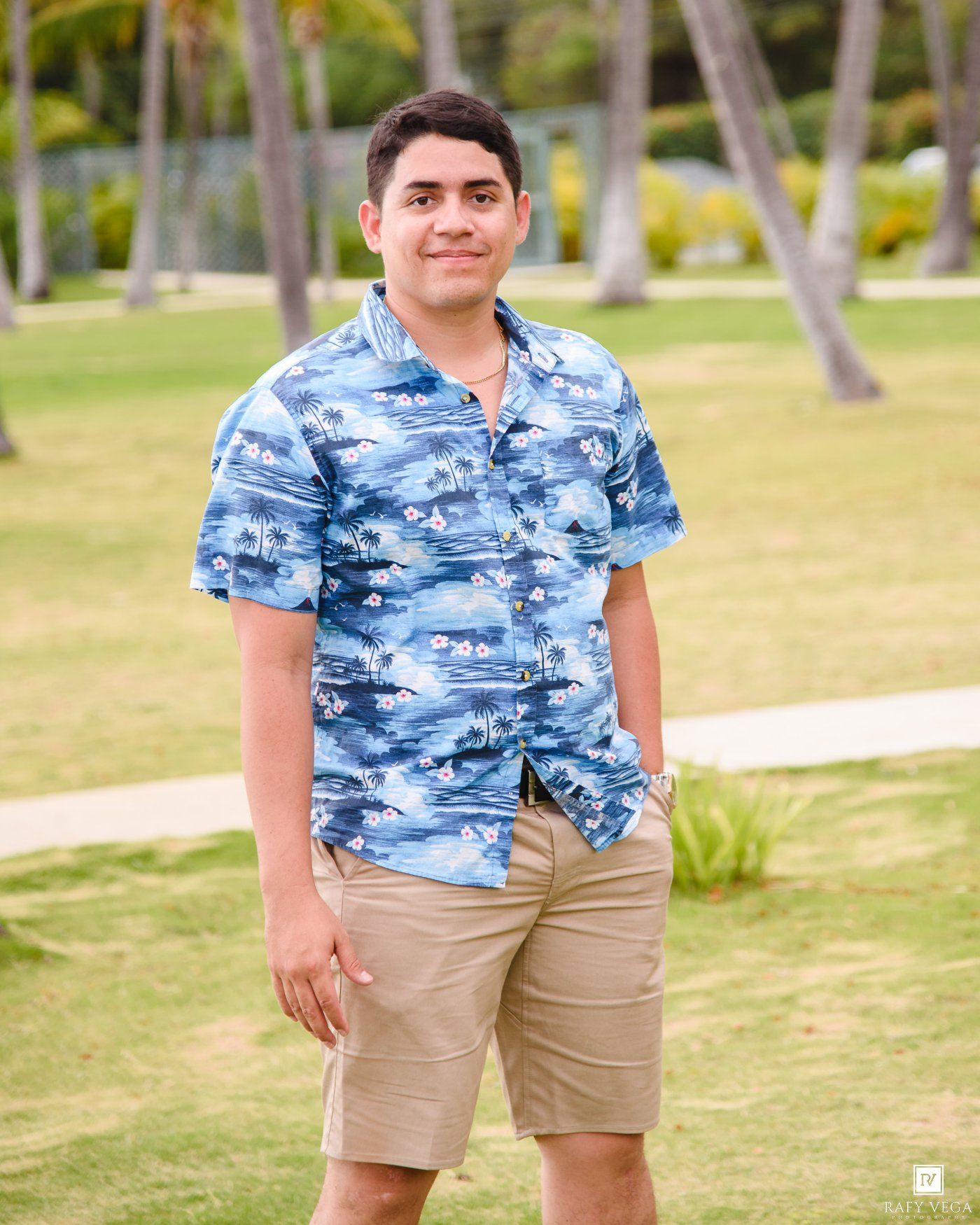 Young man in blue Hawaiian shirt and khaki shorts smiling outdoors on green lawn with palm trees.