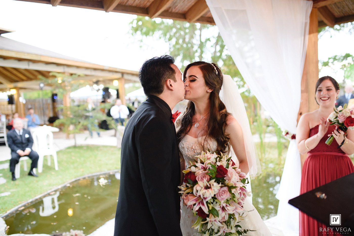 Bride and groom share first kiss at altar as bridesmaid in red dress applauds during outdoor wedding ceremony.