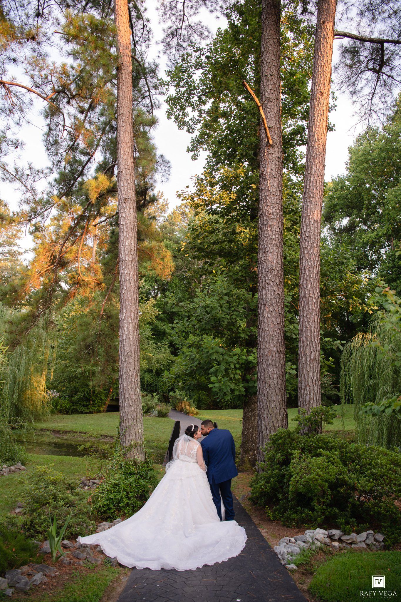 Bride and groom stand on a garden path surrounded by tall trees in a lush green park setting.