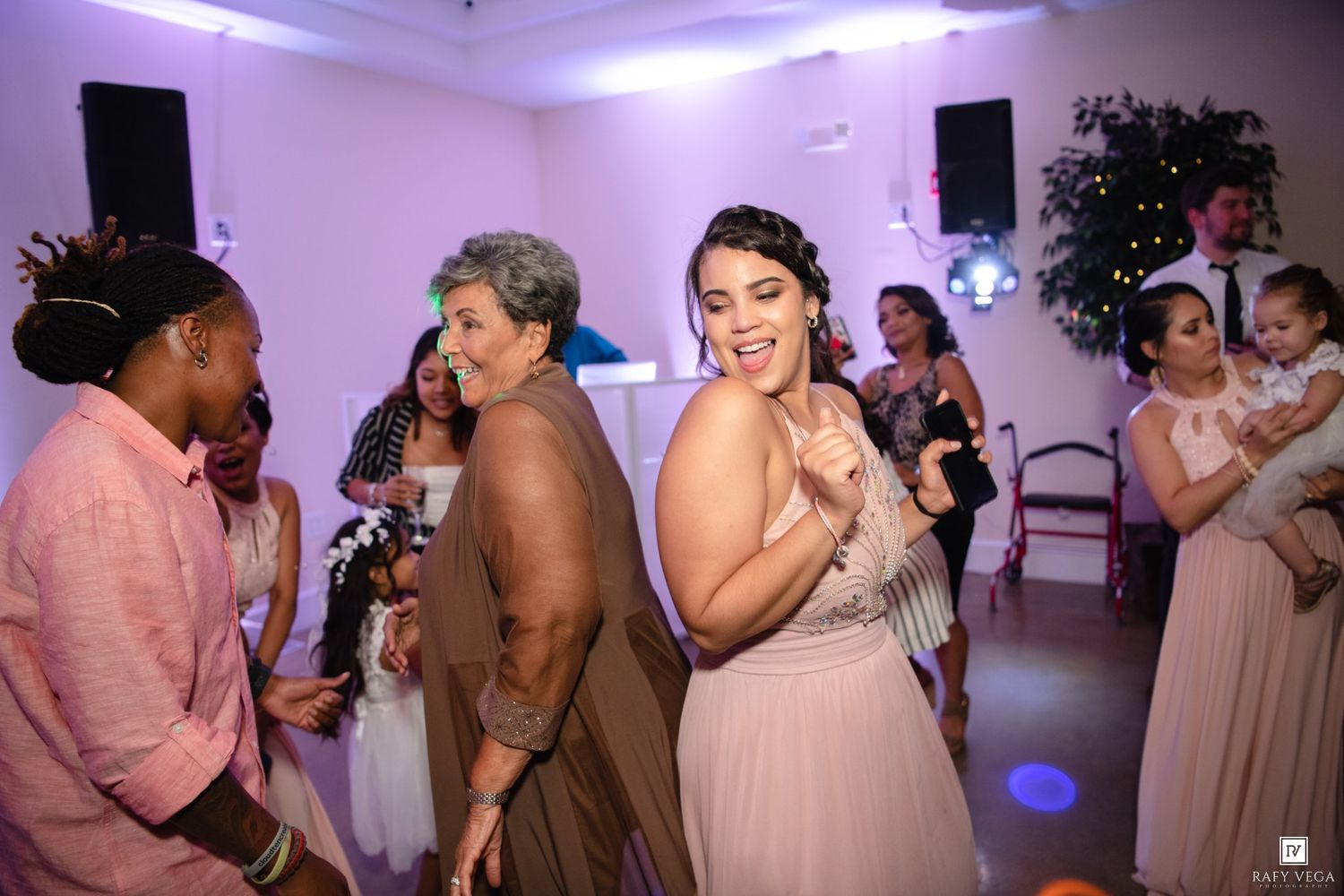 Guests dancing joyfully at a wedding reception in a decorated venue with pink lighting.