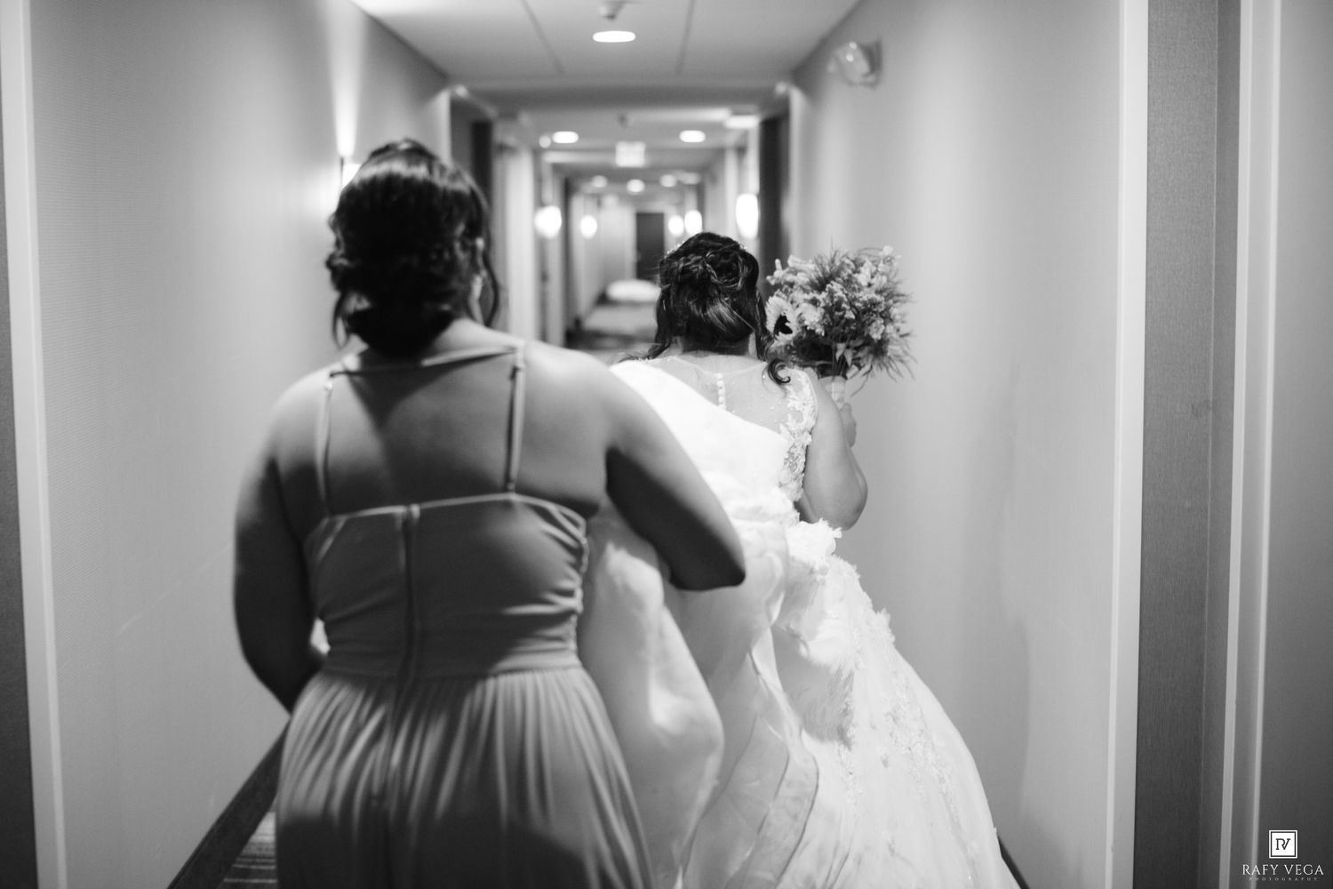 Black and white photo of a bride holding bouquet walking down hotel hallway with bridesmaid in elegant dress.