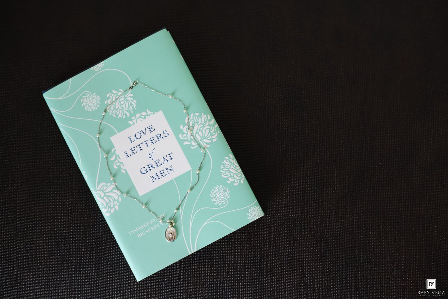 Mint green 'Love Letters of Great Men' book with delicate necklace on dark background.