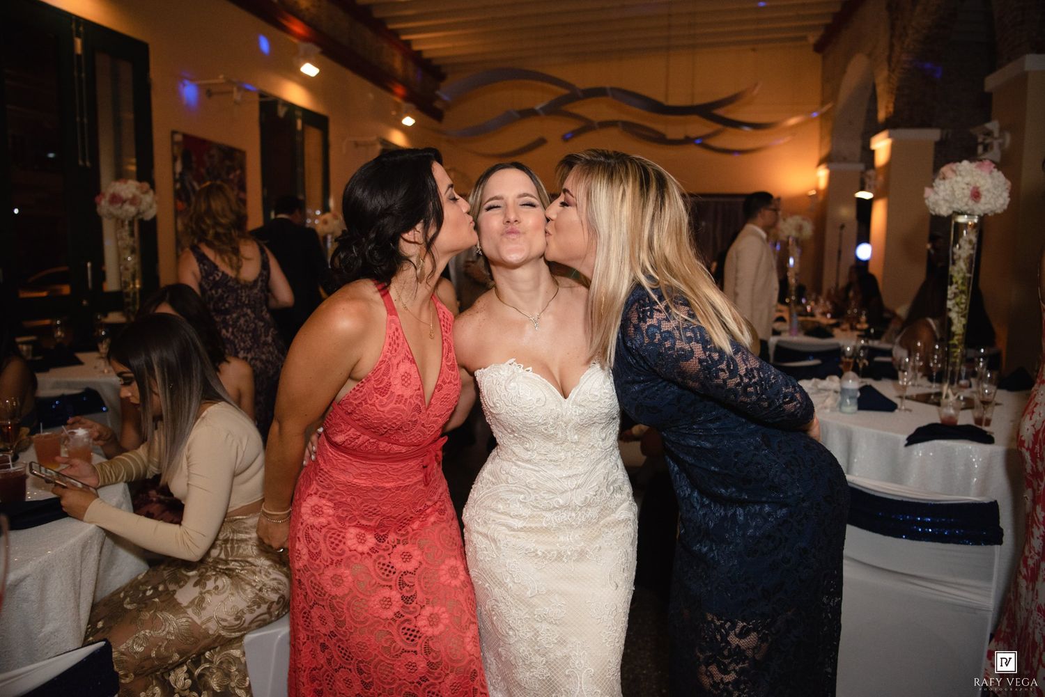 Bride in white lace gown receives kisses on cheeks from two guests in pink and blue dresses at wedding reception.