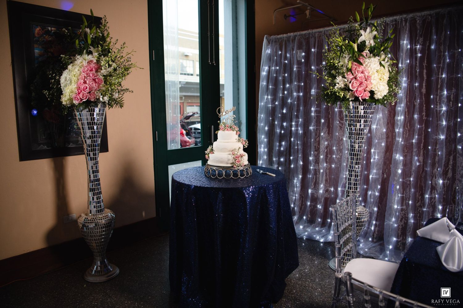 Elegant wedding cake display on navy sequin table flanked by tall floral arrangements with twinkling light backdrop.