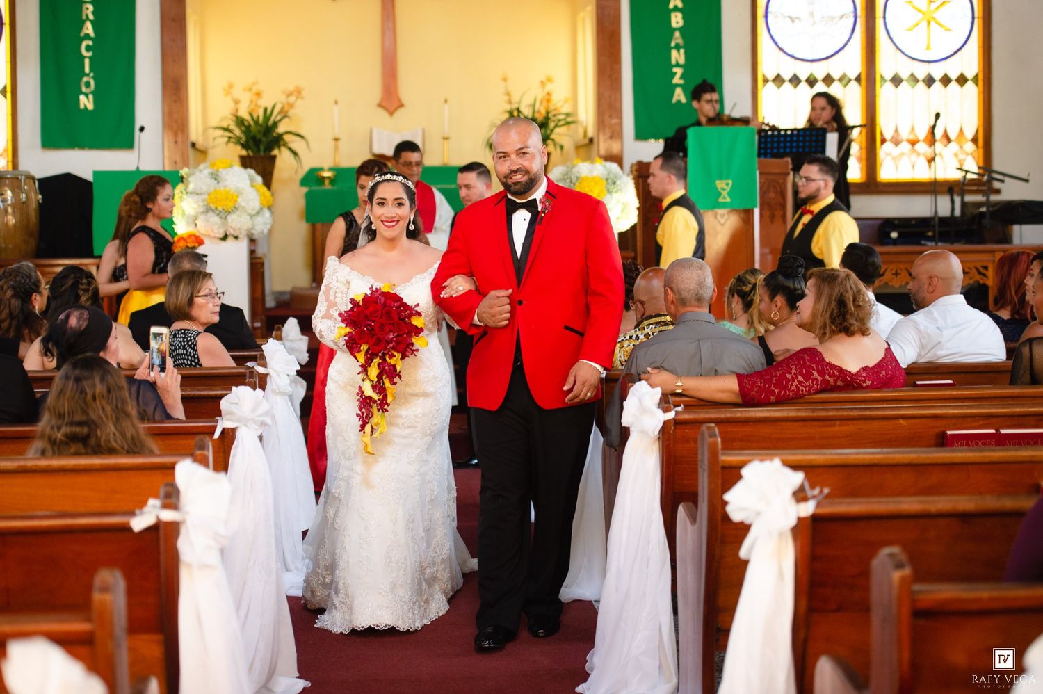Bride and groom walk down church aisle after wedding ceremony, groom in red suit, bride in white gown with red bouquet.