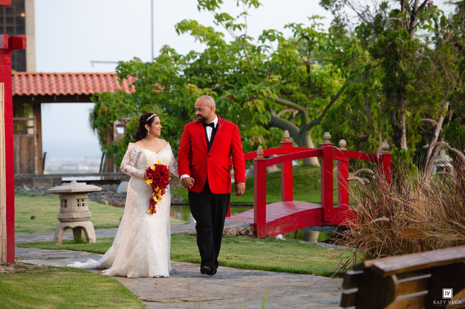 Bride in white gown and groom in red jacket walk hand in hand in a lush garden with a red bridge backdrop.