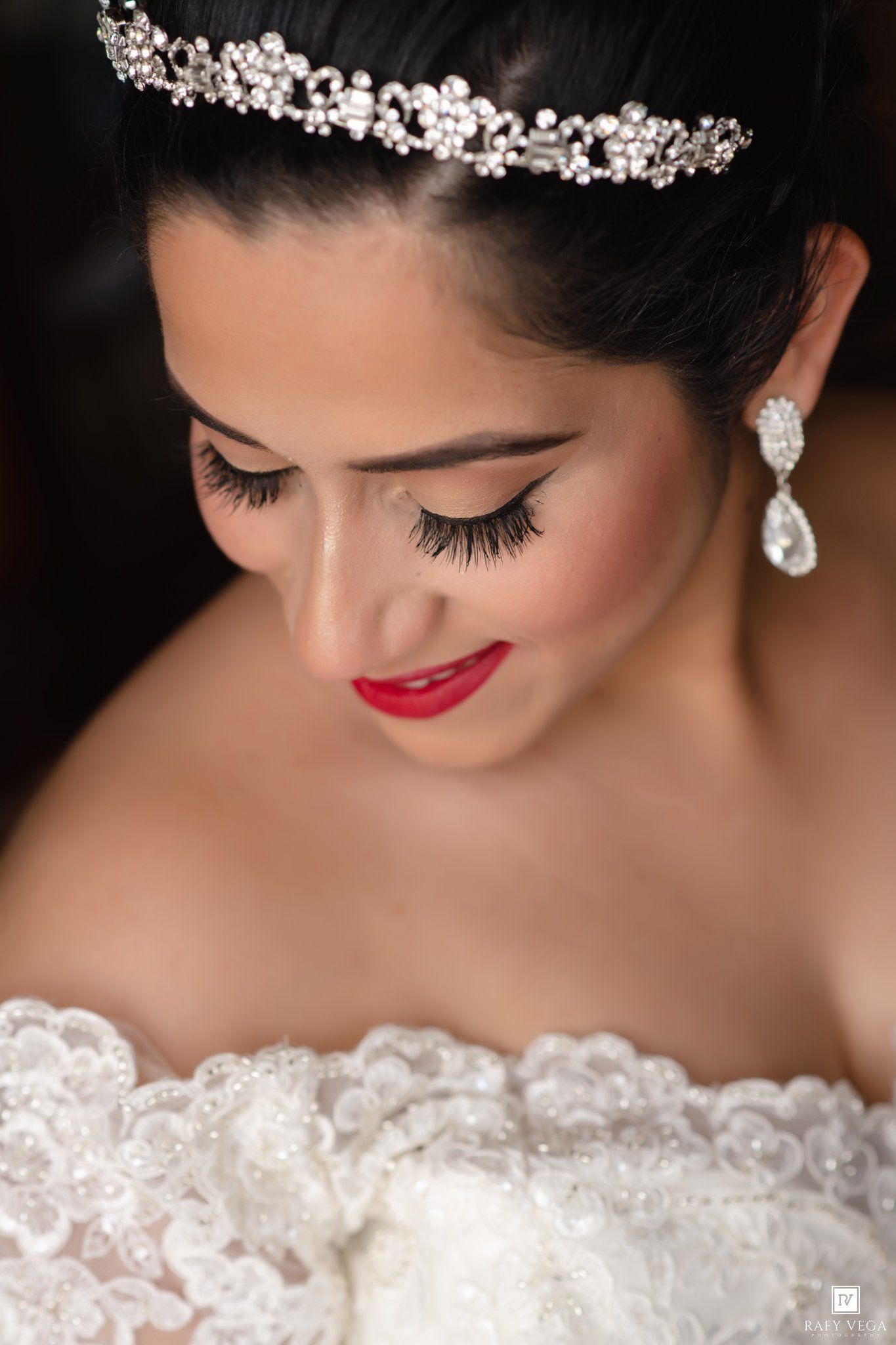Close-up of a bride wearing a crystal tiara, teardrop earrings, bold red lips, and dramatic lashes in a white lace gown.