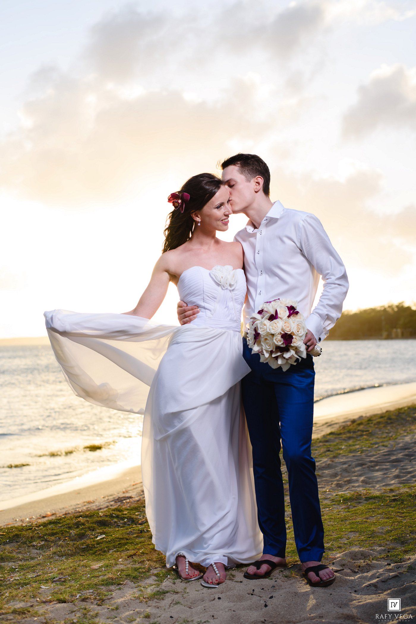 Couple in wedding attire sharing a romantic kiss on a tropical beach at sunset, bride holding bouquet.