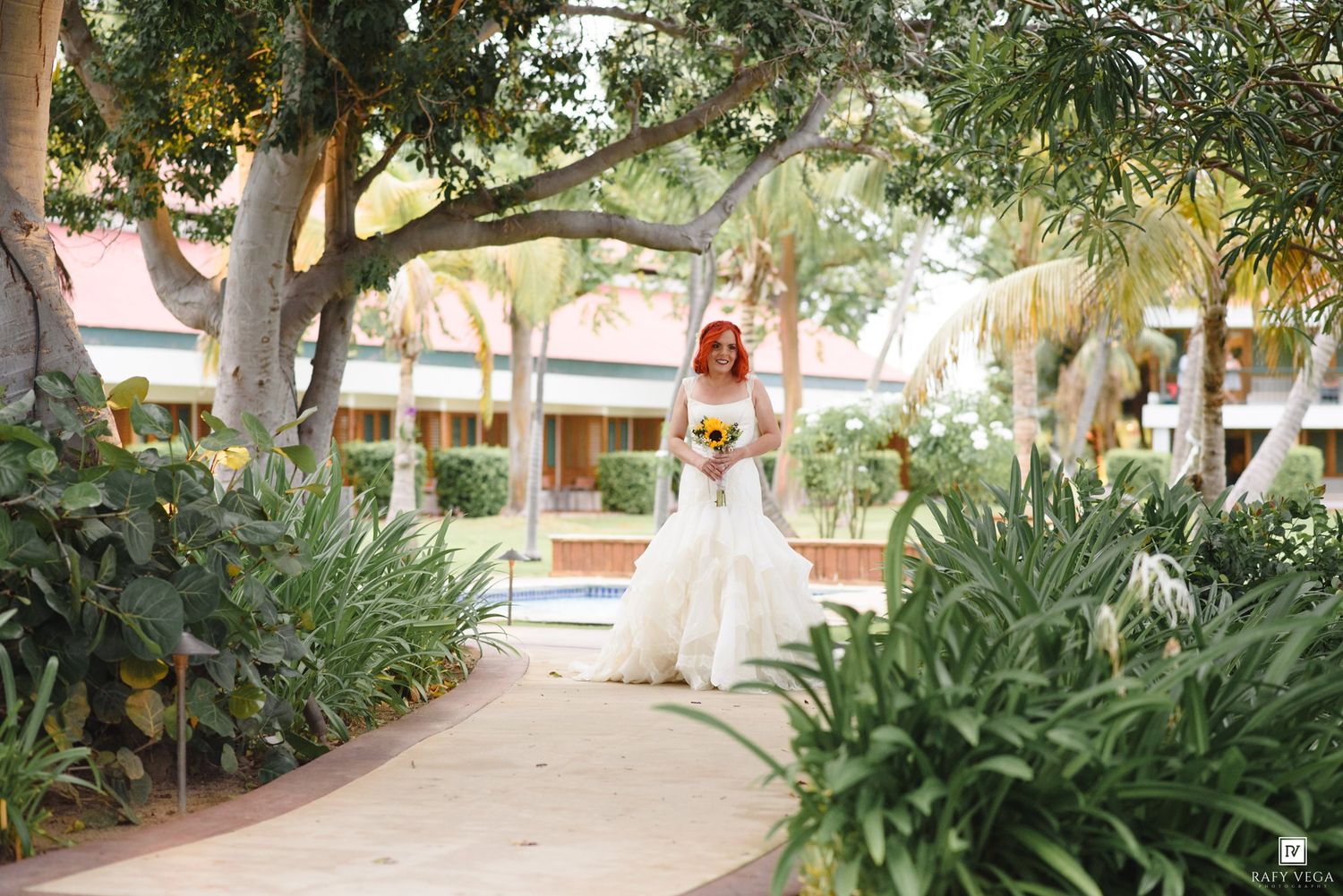 Bride in white gown holding yellow bouquet stands on garden path surrounded by tropical trees and lush greenery.