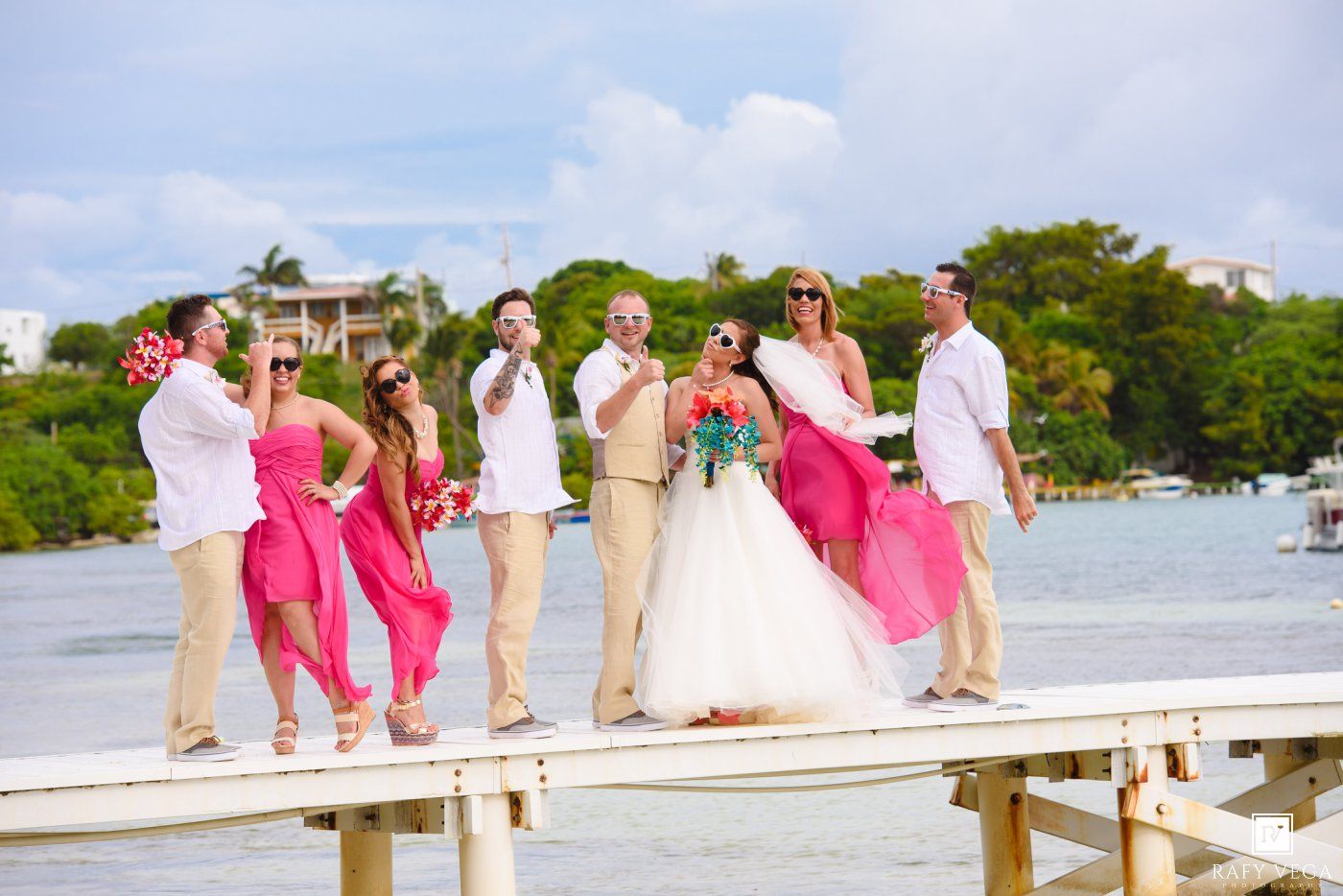 Wedding party posing on a wooden dock by the water, bridesmaids in pink dresses, groomsmen in khaki, tropical setting.