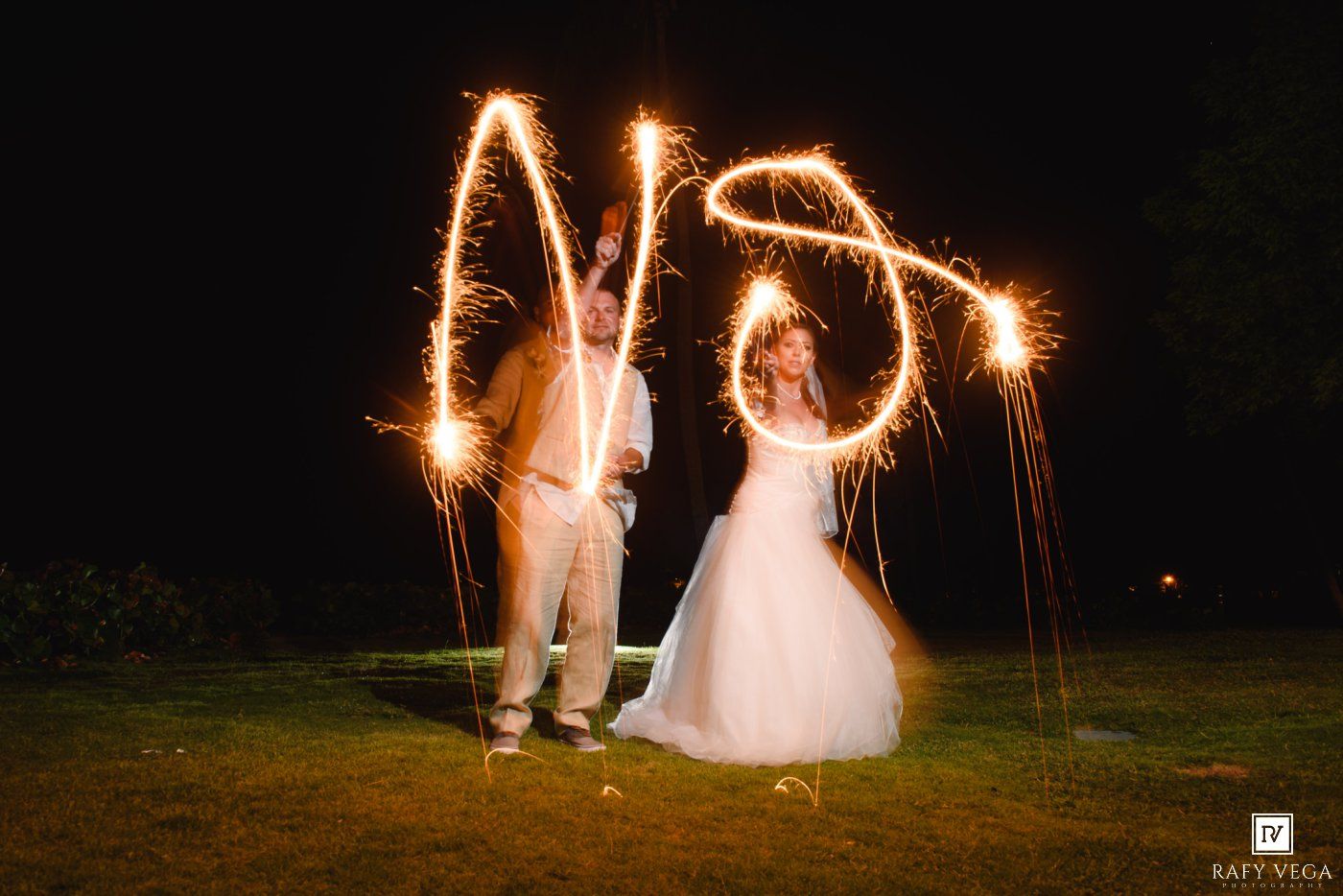 Bride and groom writing initials with sparklers at night during wedding celebration outdoors.
