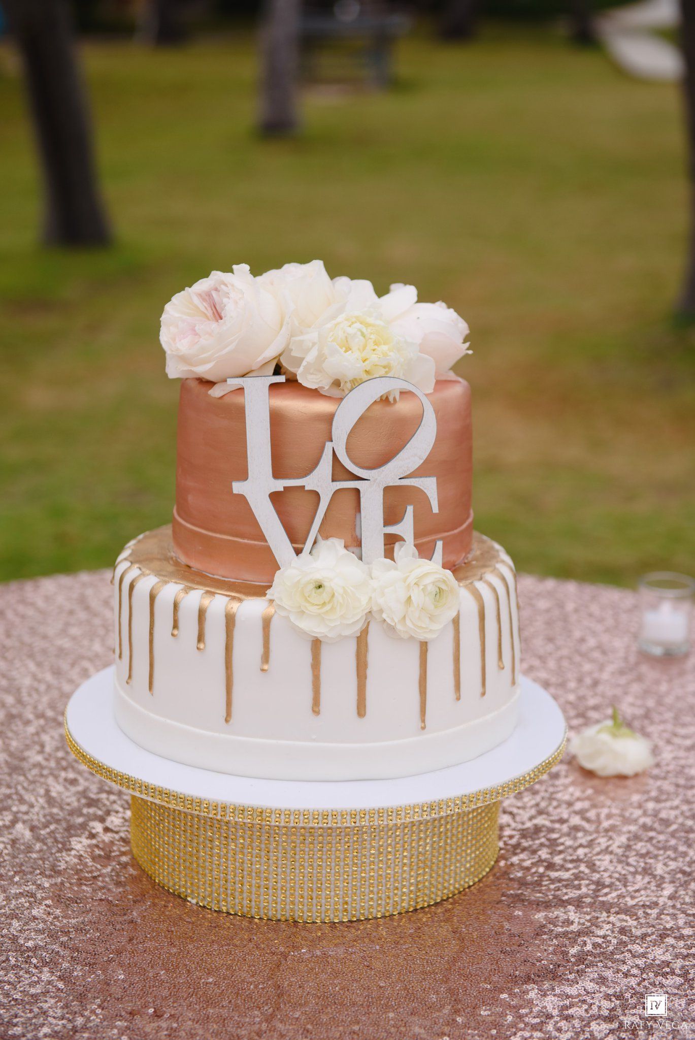 Two-tier rose gold and white wedding cake with LOVE topper, gold drip details, and white floral decorations on sequin stand.