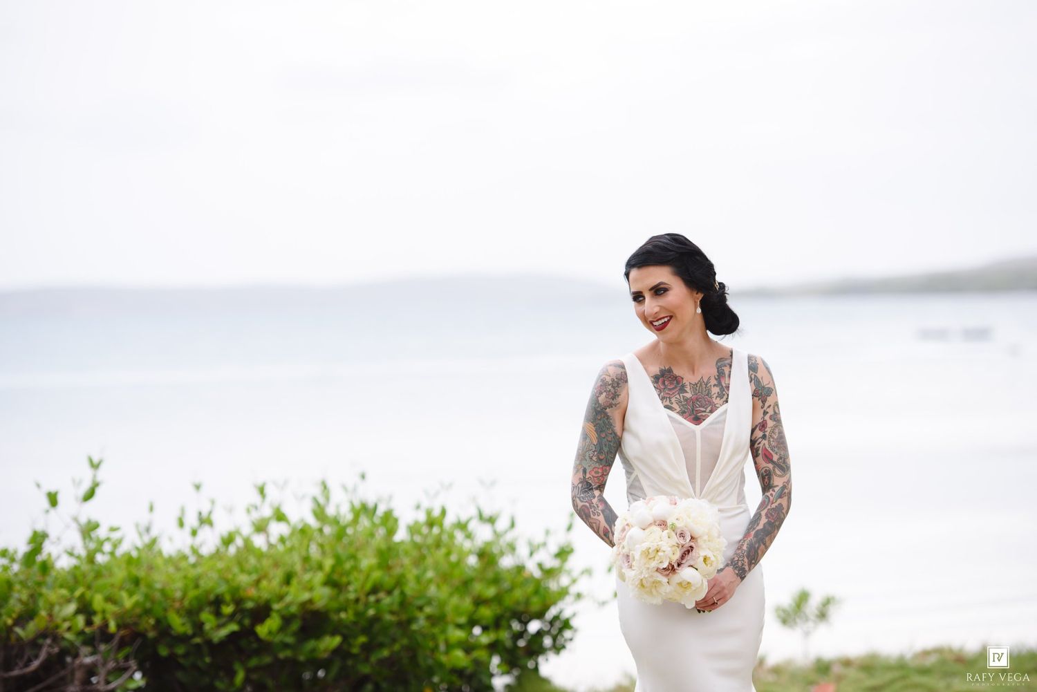 Tattooed bride in sleek white gown holding white bouquet smiling by ocean shore.