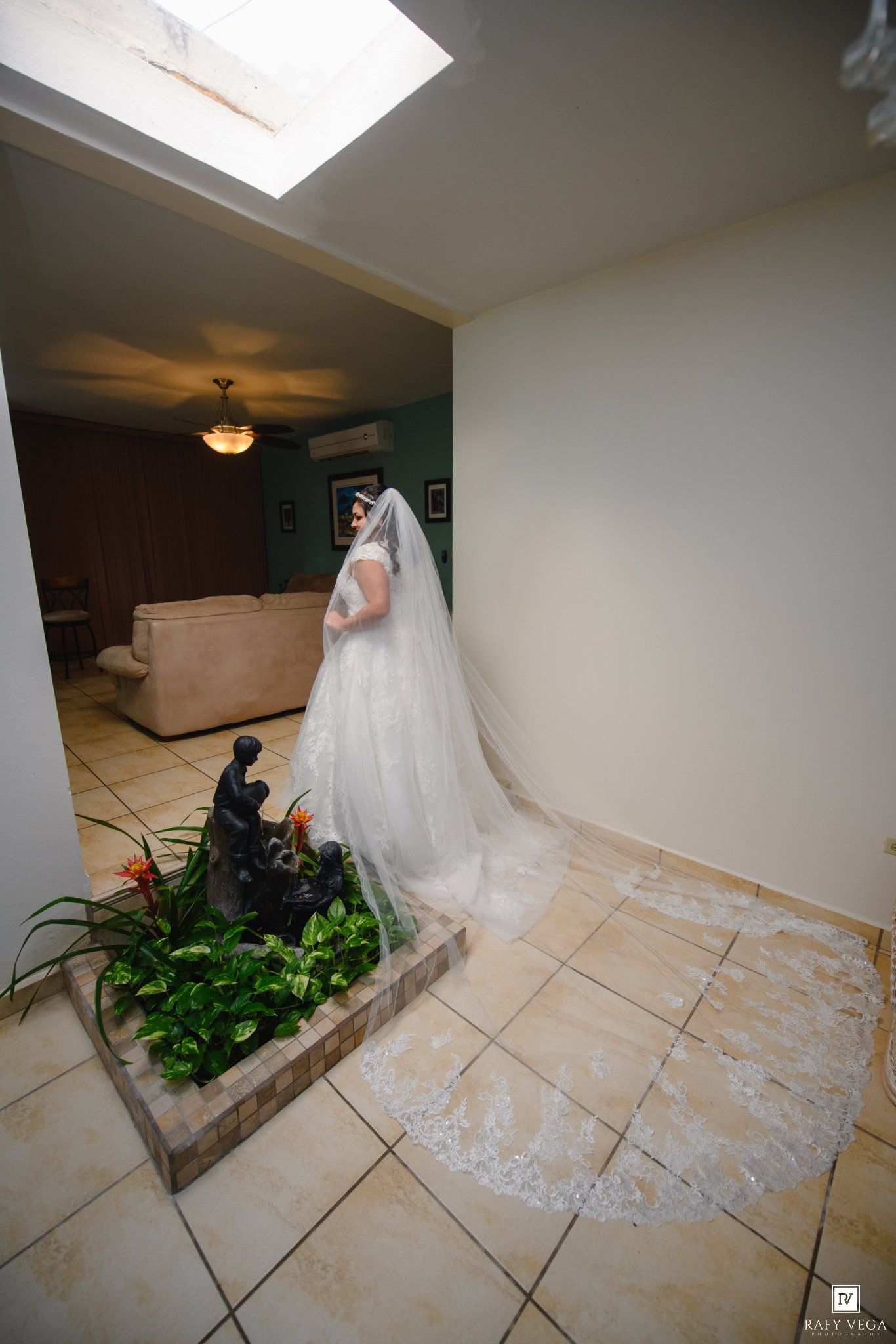 Bride in white wedding gown and long lace veil standing near indoor garden statue in tiled hallway.