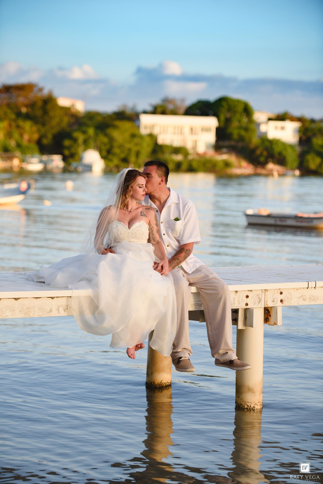 Bride and groom share a romantic kiss on a waterfront dock, with lush greenery and calm waters in the background.