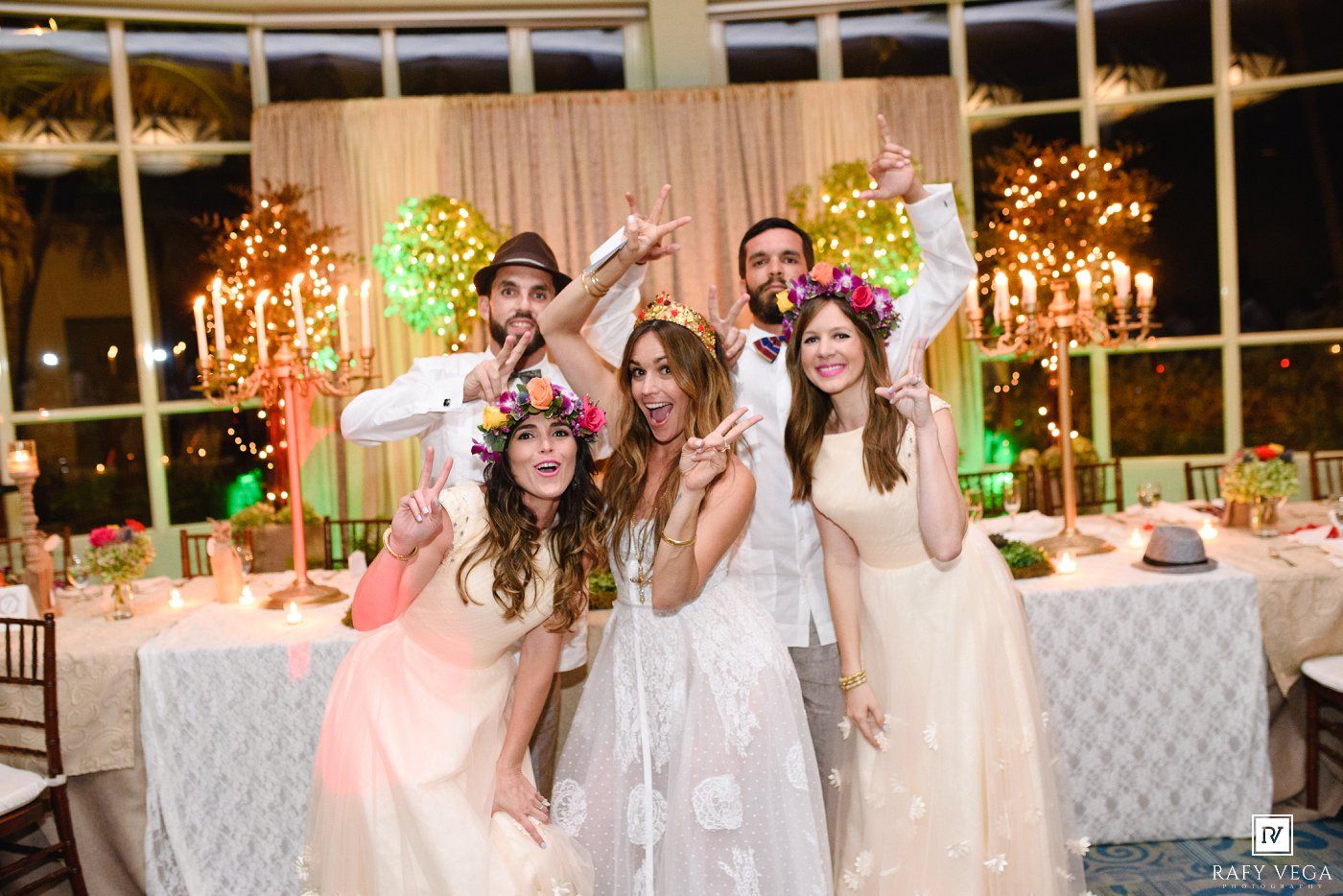 Wedding party poses joyfully at festive reception with floral crowns, candlelit decor, and colorful arrangements.