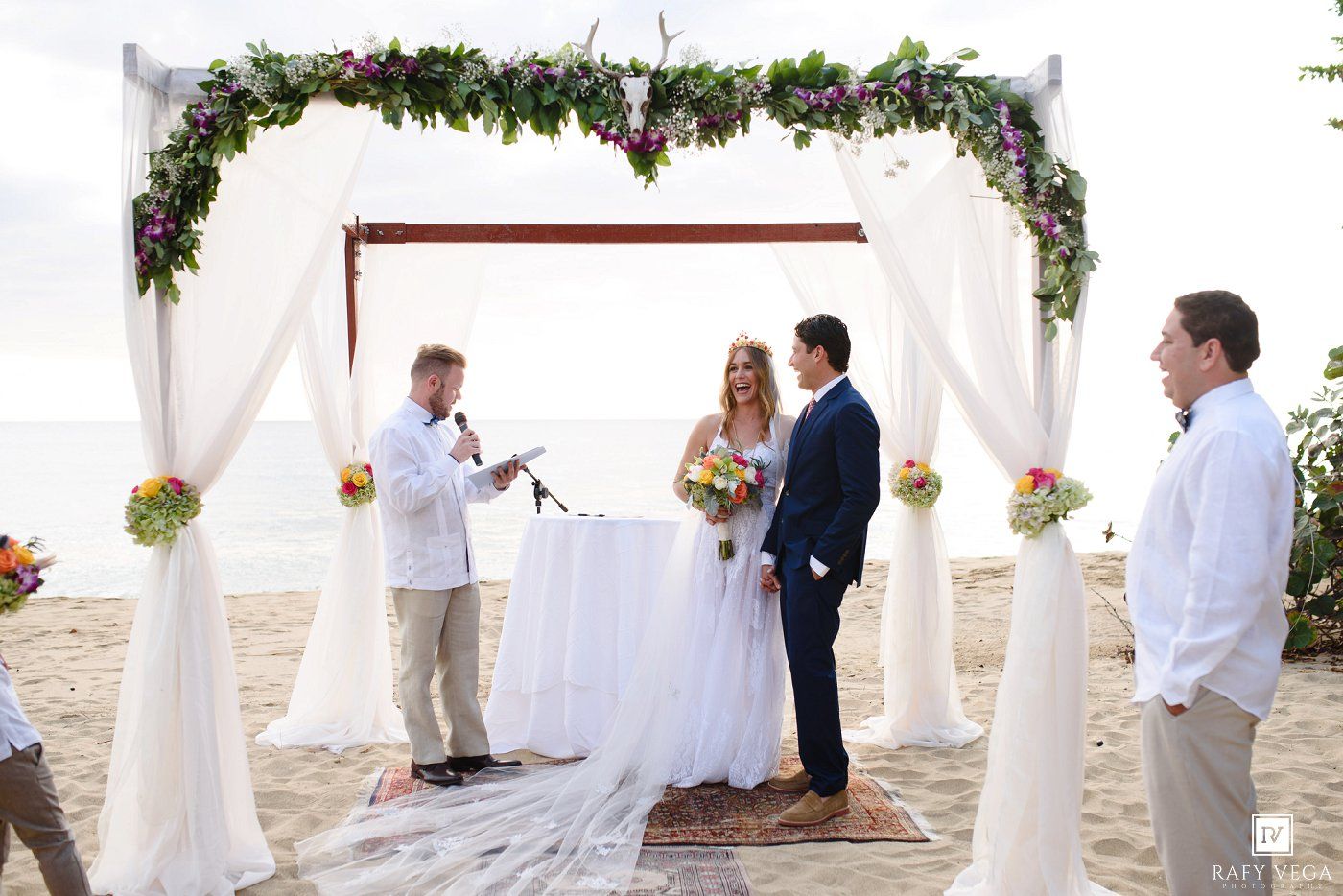 Couple exchanges vows at a romantic beach wedding ceremony under a floral arch adorned with white draping.