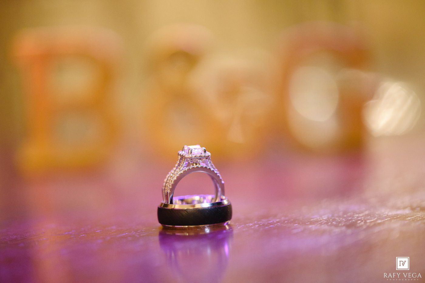 Wedding rings with diamond engagement ring displayed on purple surface with blurred B&G monogram letters in gold background.