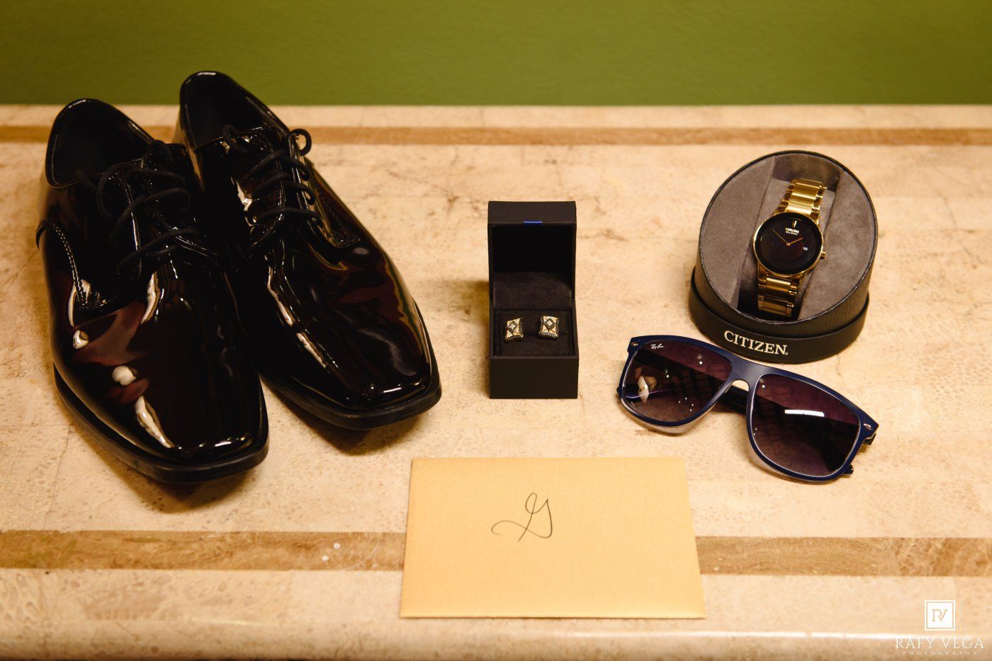Black patent leather dress shoes, cufflinks, Citizen watch, sunglasses, and an envelope arranged on a shelf.