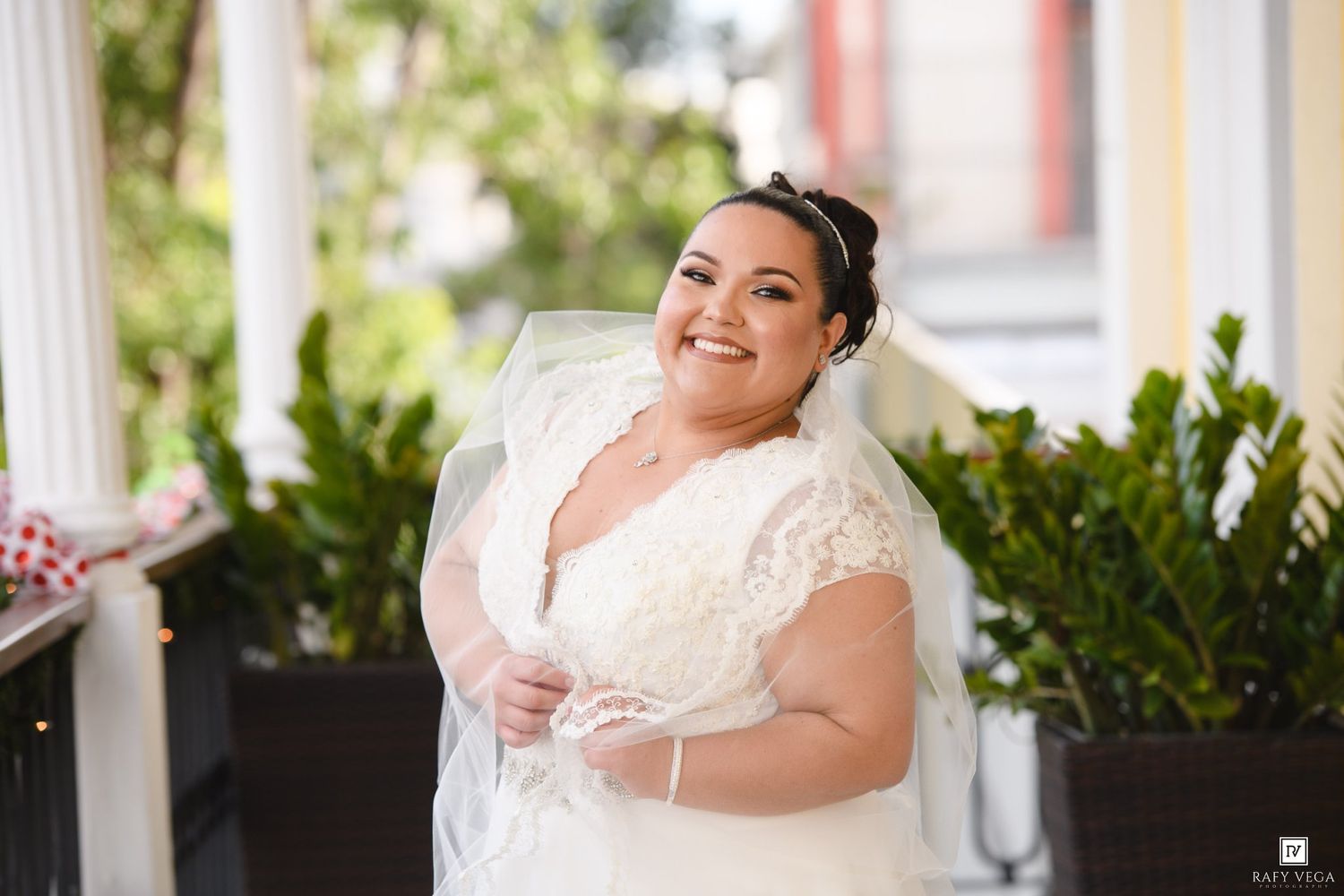 Smiling bride in white lace wedding dress and veil poses outdoors on a porch surrounded by greenery.
