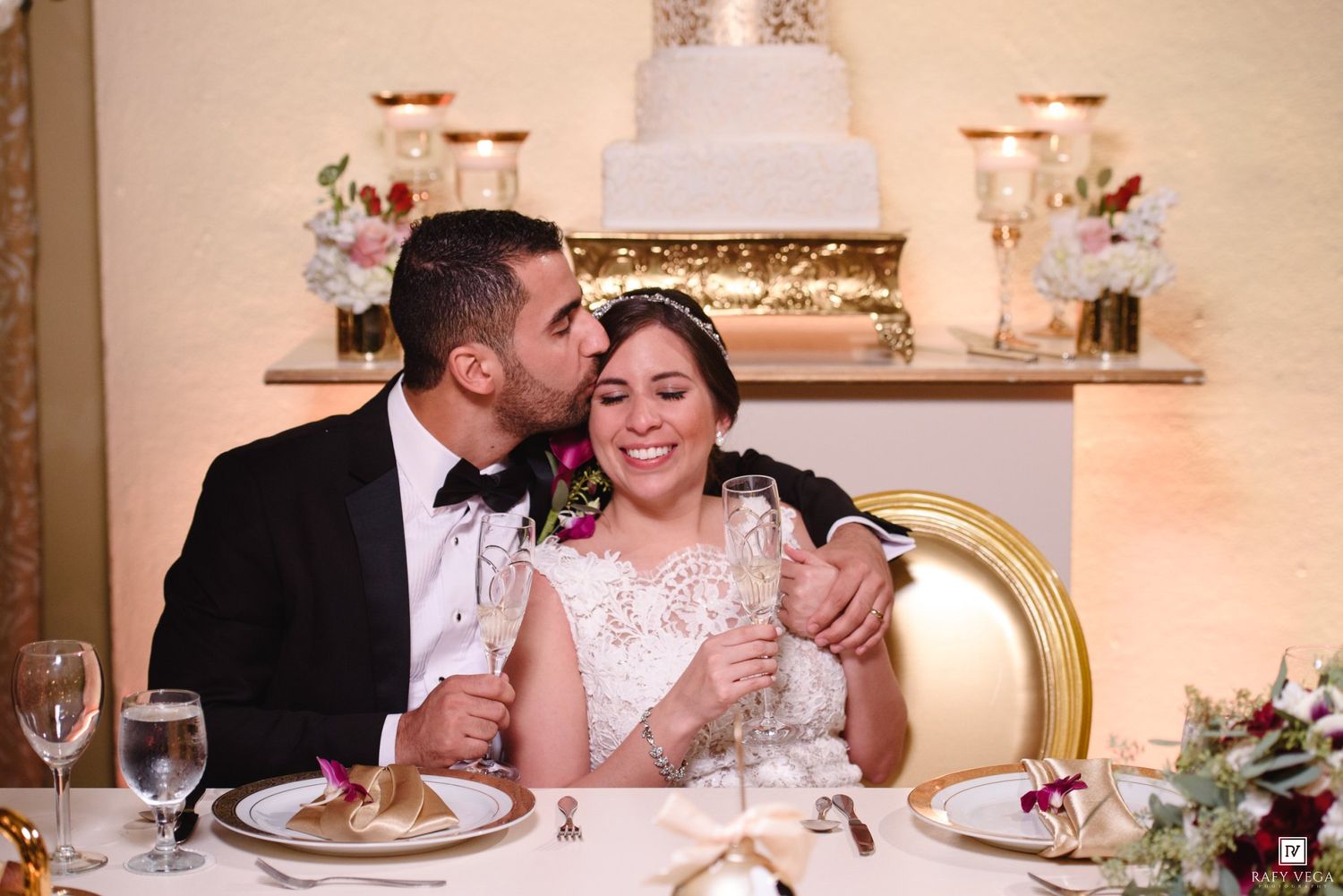 Bride and groom share a romantic moment at wedding reception sweetheart table with white cake in background.