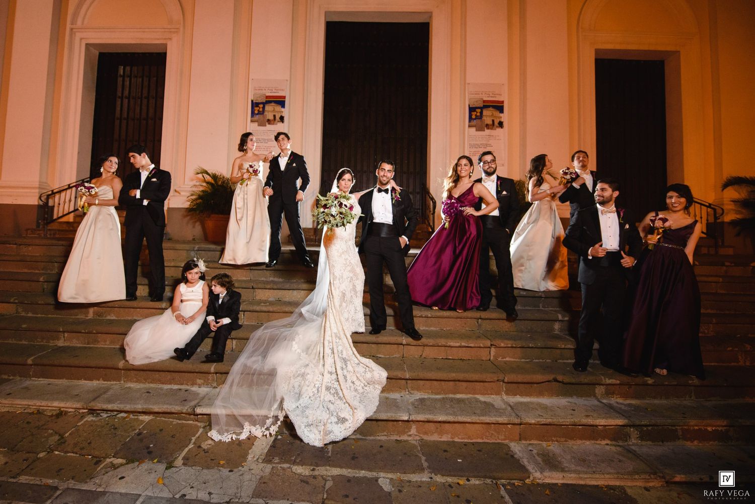 Wedding party poses on grand building steps, bride in lace gown with long veil, groom in tuxedo, bridesmaids in burgundy.
