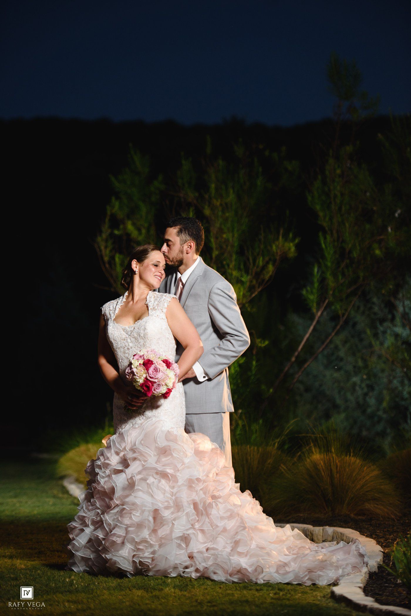 Bride in ruffled white gown holds pink bouquet as groom in gray suit embraces her during romantic nighttime outdoor wedding portrait.