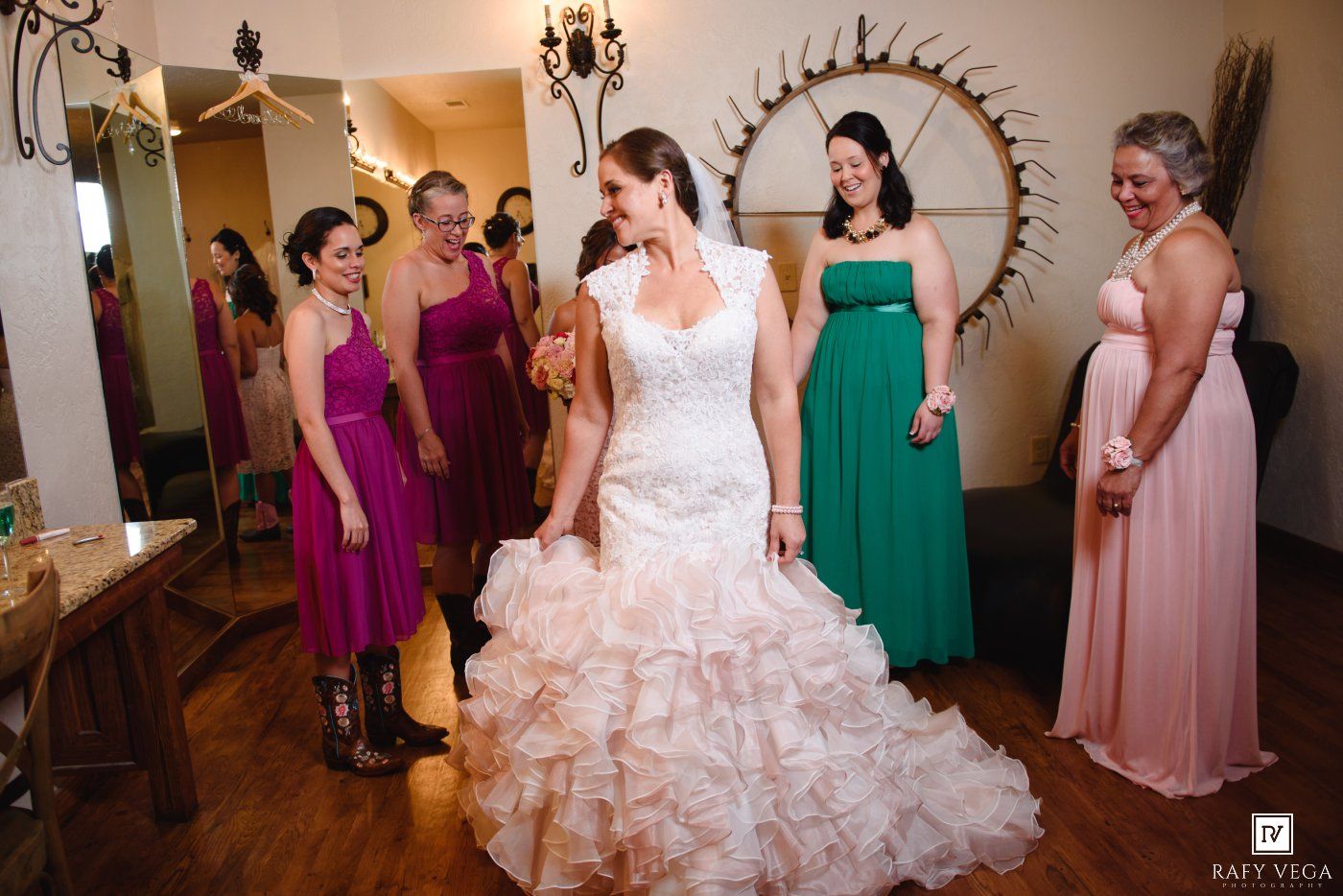 Bride in white ruffled gown surrounded by bridesmaids in colorful dresses during wedding preparations indoors.