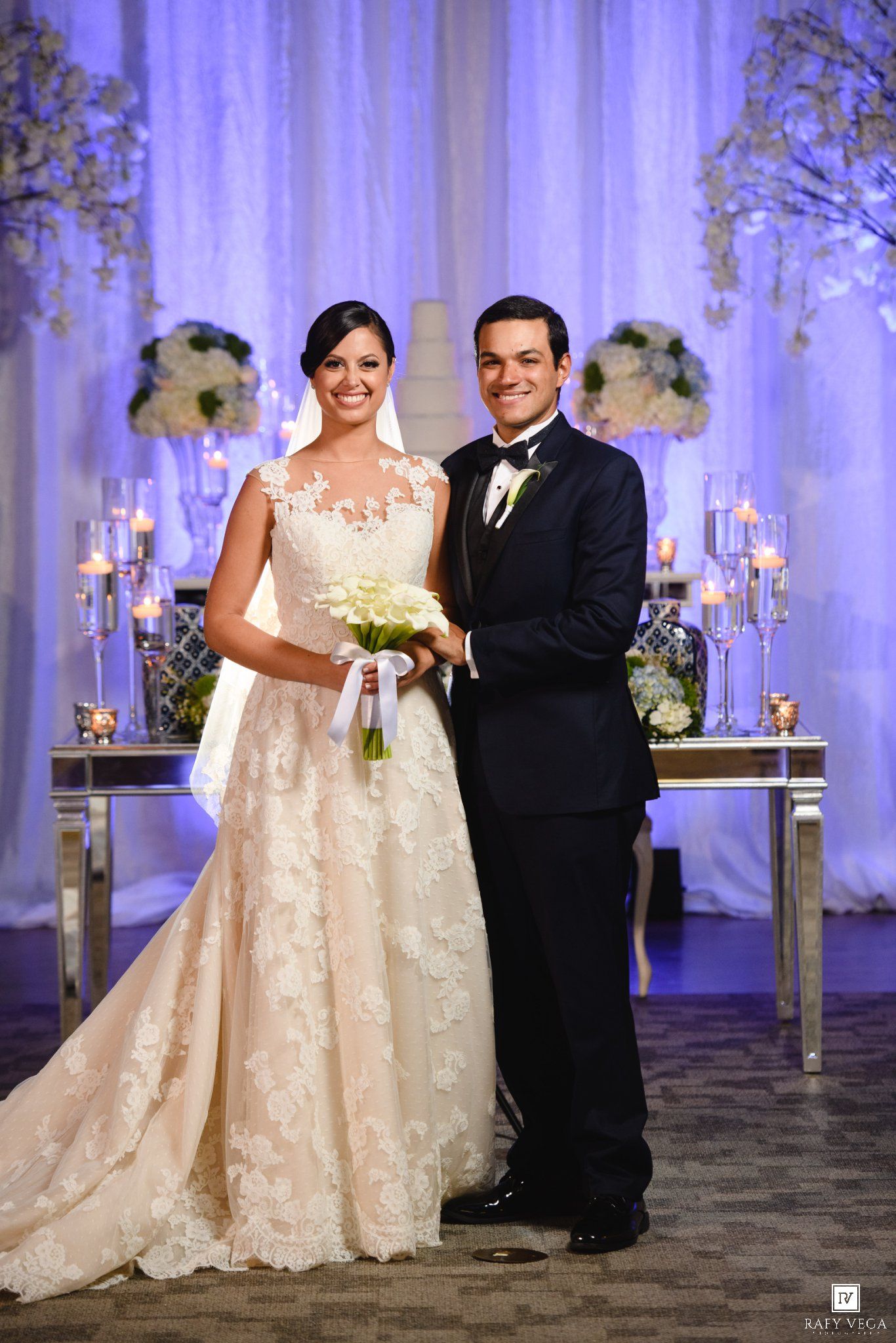 Bride in lace gown holding white bouquet stands with groom in black tuxedo at elegant wedding reception.