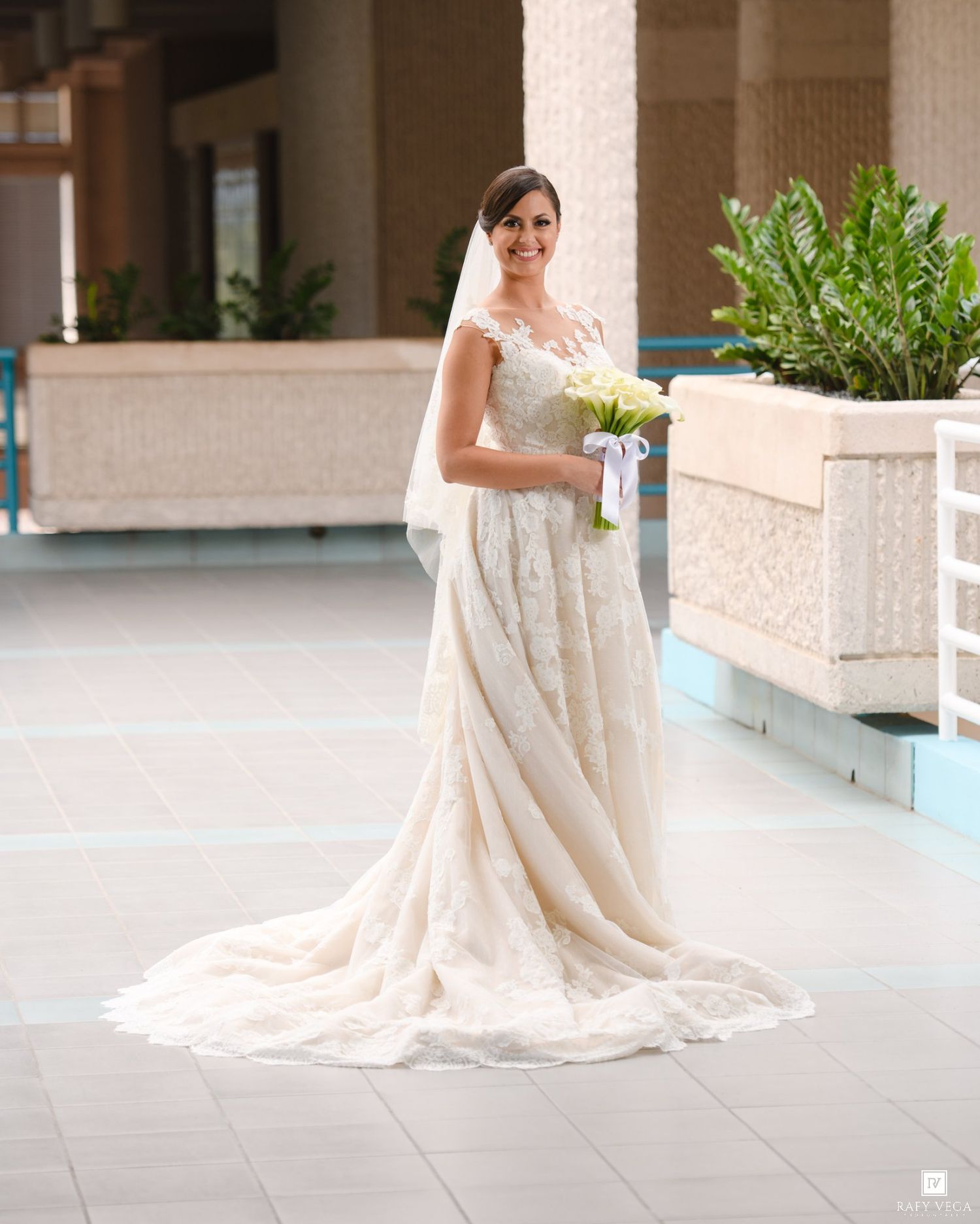 Bride in lace wedding gown with veil holding white bouquet, standing in elegant venue with tiled floors.