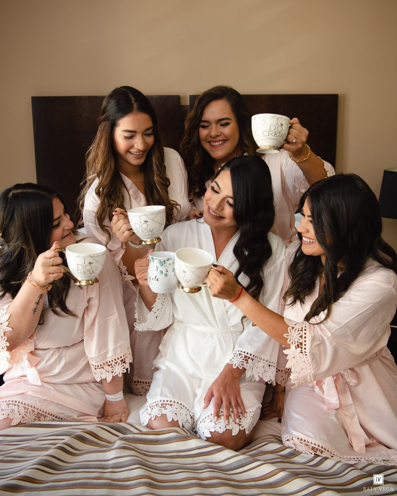 Bride in white robe toasting mugs with bridesmaids in pink robes on a bed during wedding morning preparations.