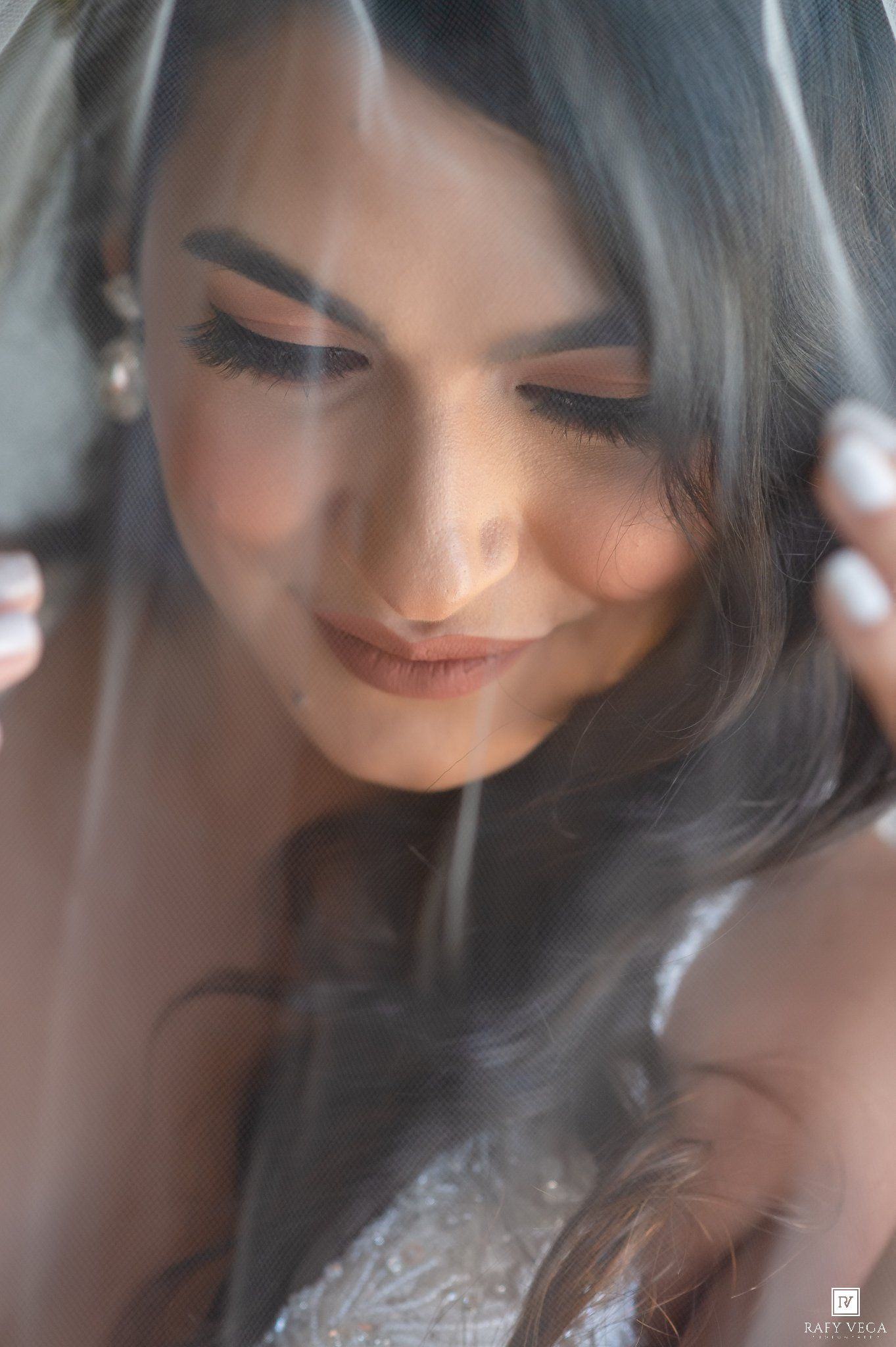 Bride smiling softly under a sheer veil, wearing lace gown with elegant makeup and loose waves.