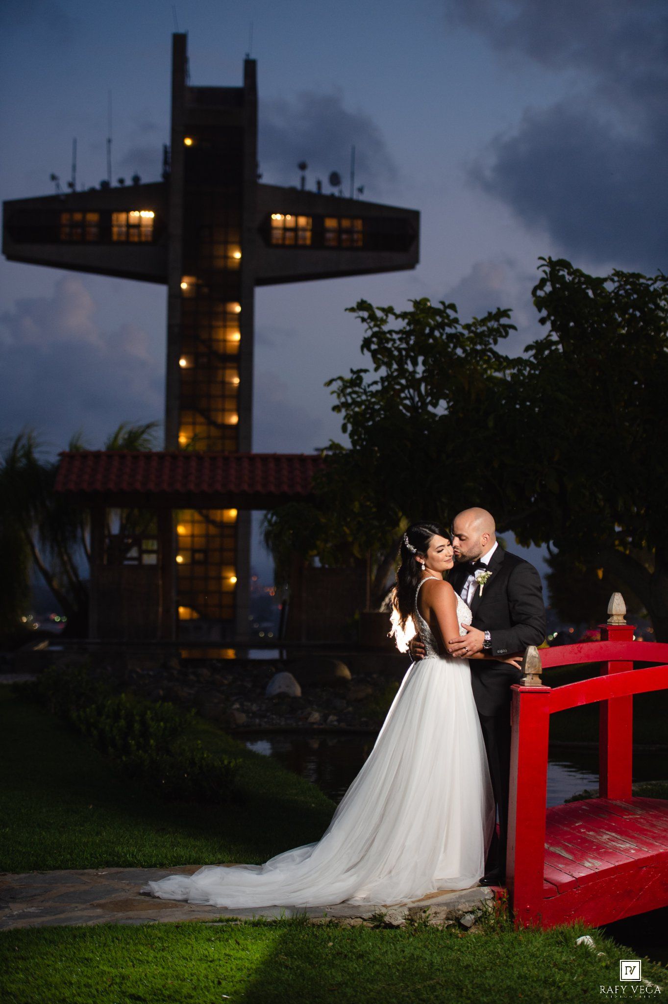 Bride and groom share romantic kiss at dusk with illuminated cross tower backdrop at outdoor wedding venue.