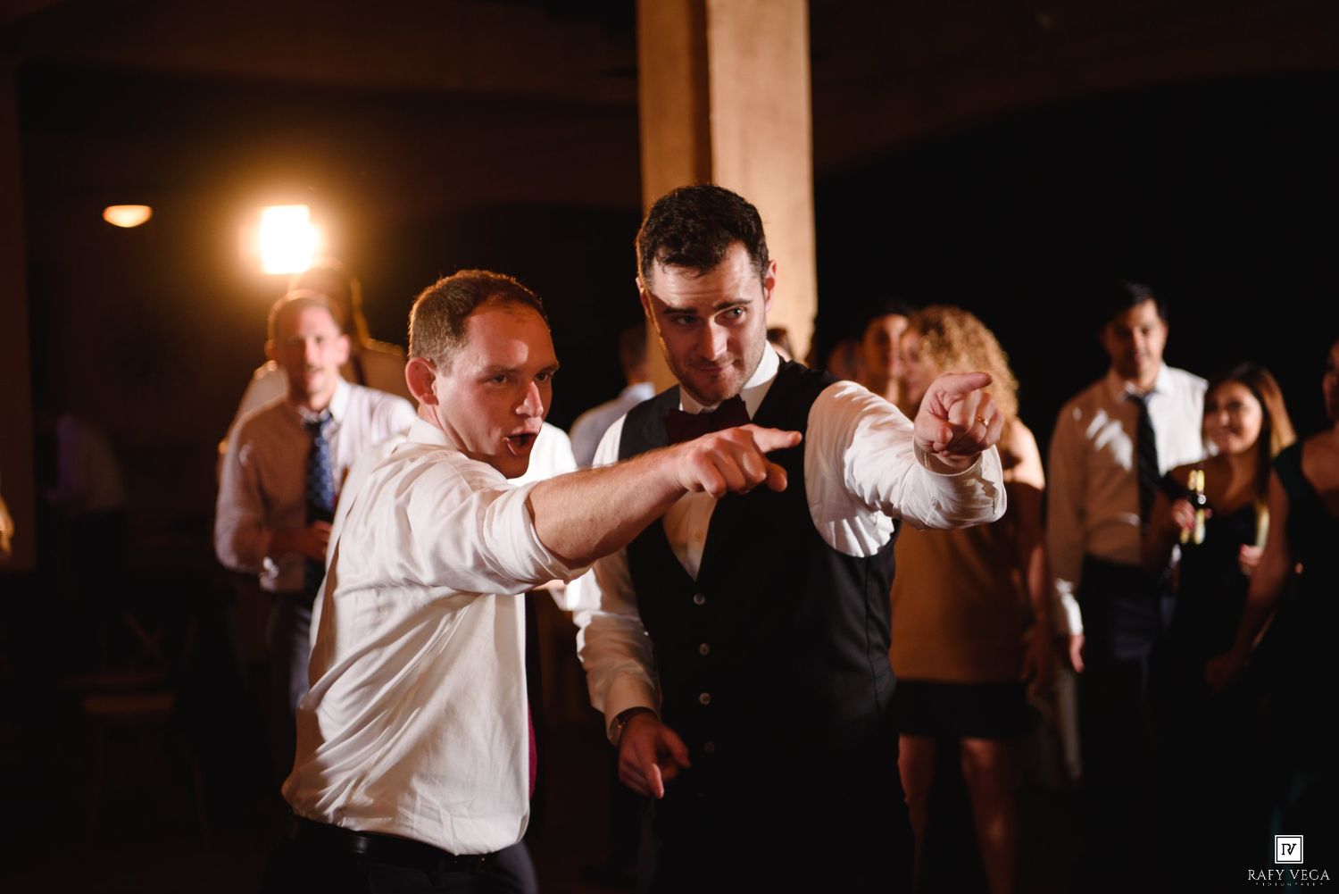Two men dancing energetically at a wedding reception, pointing forward on a dimly lit dance floor.
